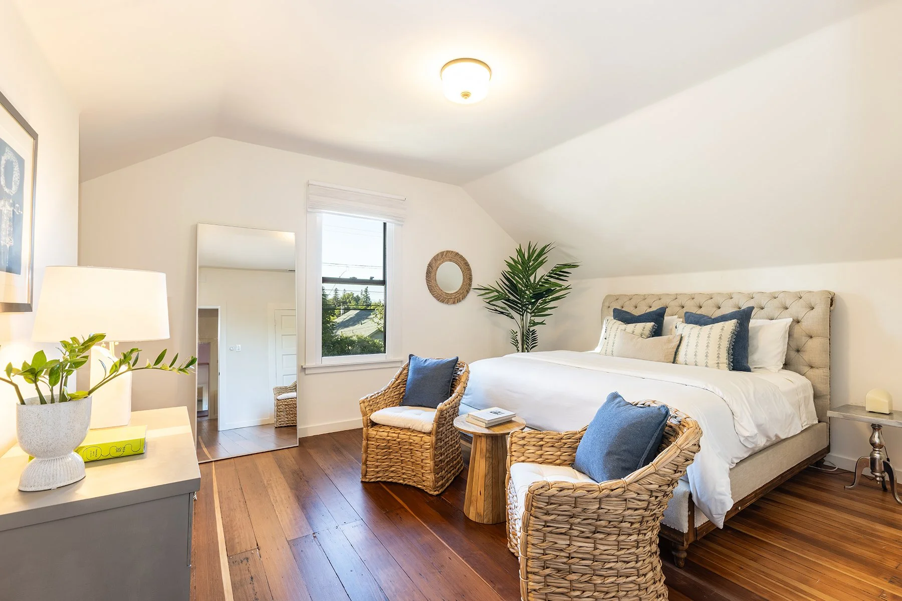 Bright bedroom with white walls, a beige headboard, and blue and white pillows on a white bed. Wicker chairs with blue pillows, a small wooden side table, a potted plant, a mirror, a window with white blinds, and a ceiling light.