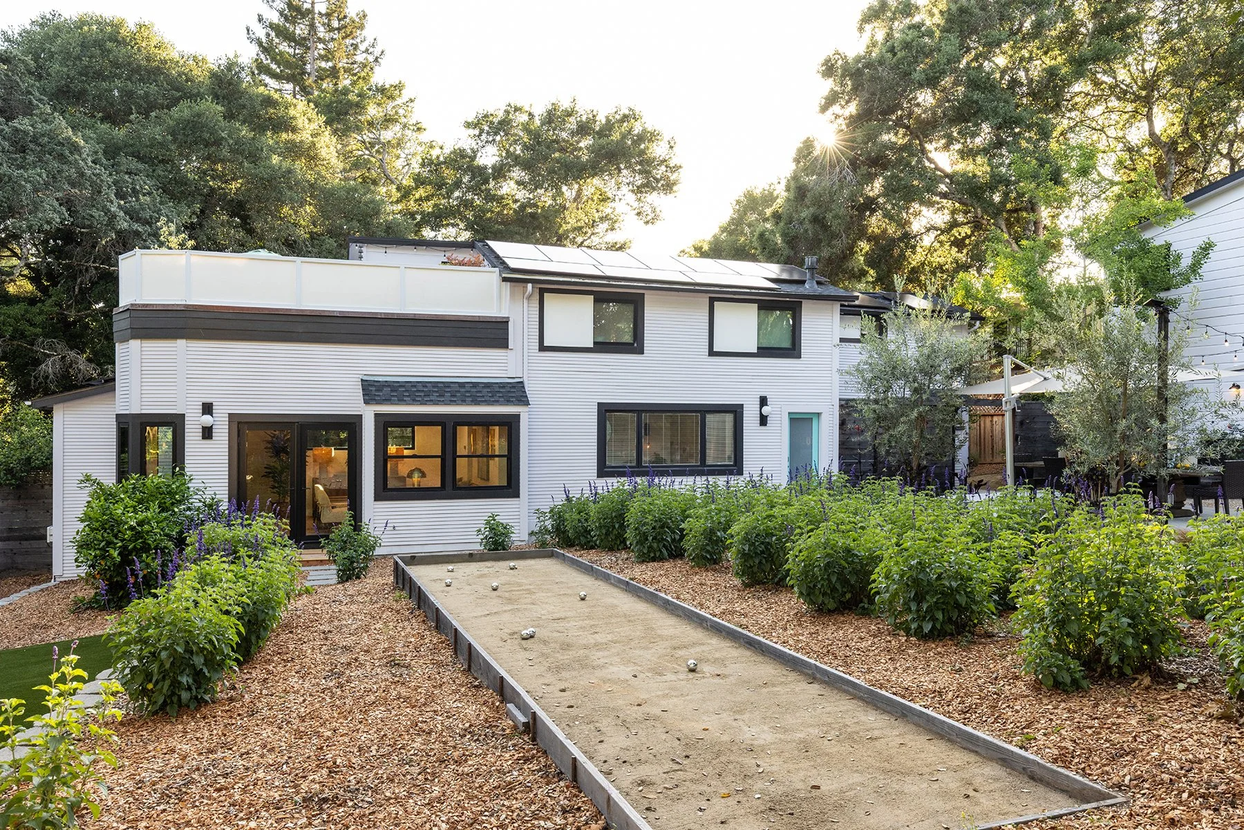 A modern two-story house with white siding and black trim, surrounded by green trees and a landscaped yard, featuring a bocce ball court with metal balls, a garden with purple flowers, and outdoor seating areas at sunset.