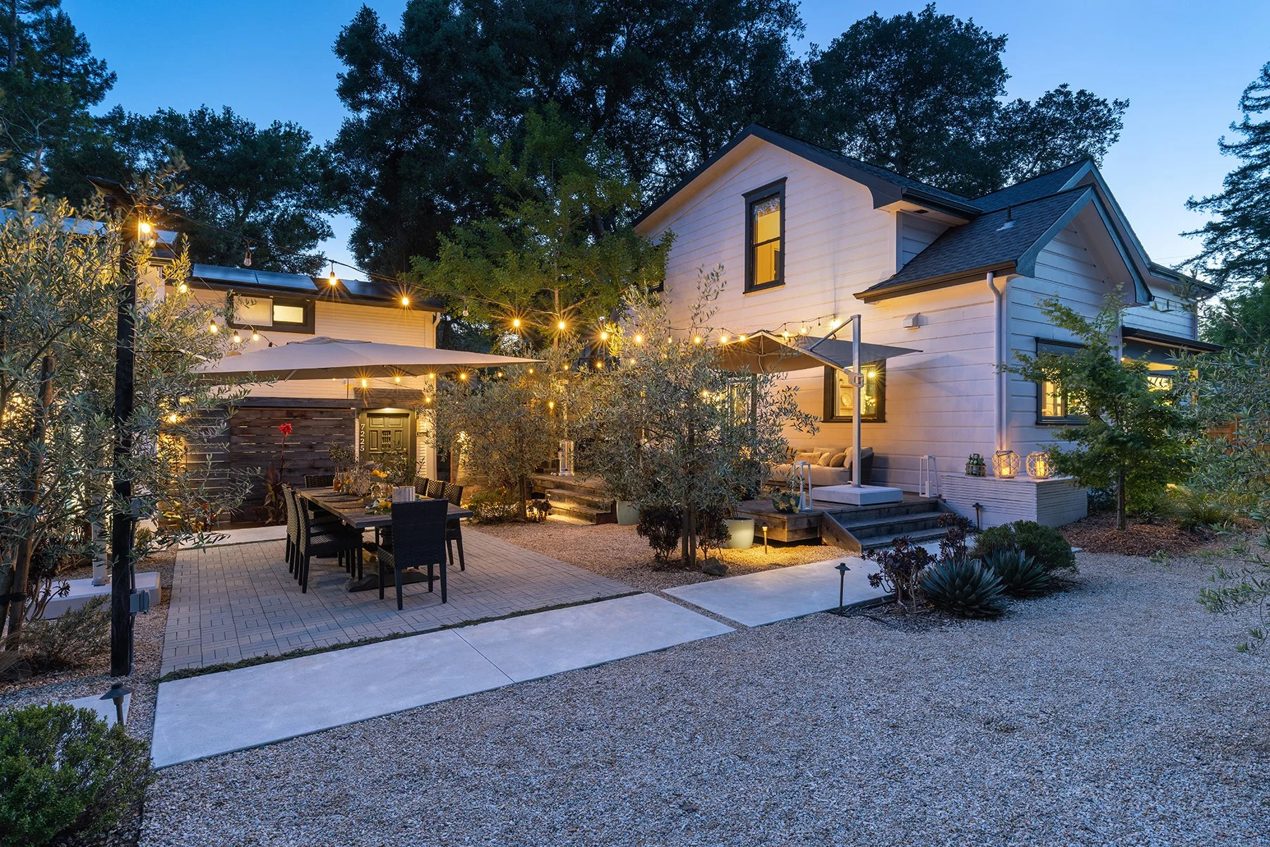 A backyard patio area of a house illuminated with string lights at dusk, featuring a dining table with chairs, potted plants, trees, and a cozy seating area with lanterns and outdoor furniture.