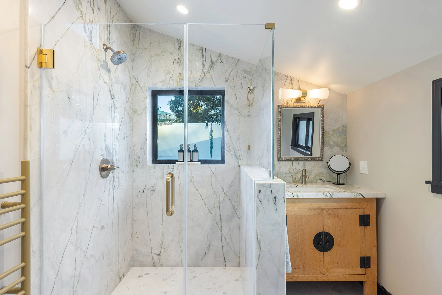 Modern bathroom with a marble shower, window, and wooden vanity with a mirror and lighting.