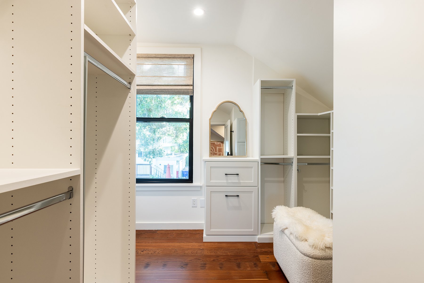 Empty walk-in closet with white built-in shelves and drawers, a window with a view of trees outside, and a fluffy white bench.
