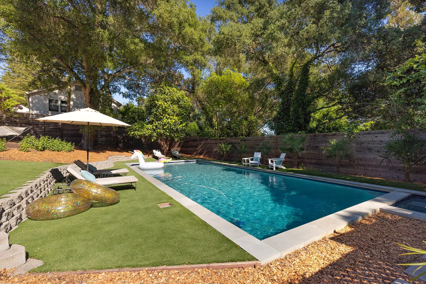 A backyard swimming pool area with lounge chairs, an umbrella, and an inflatable unicorn float. There are trees and a wooden fence in the background, and the area is decorated with gold and rainbow-colored pool floats.