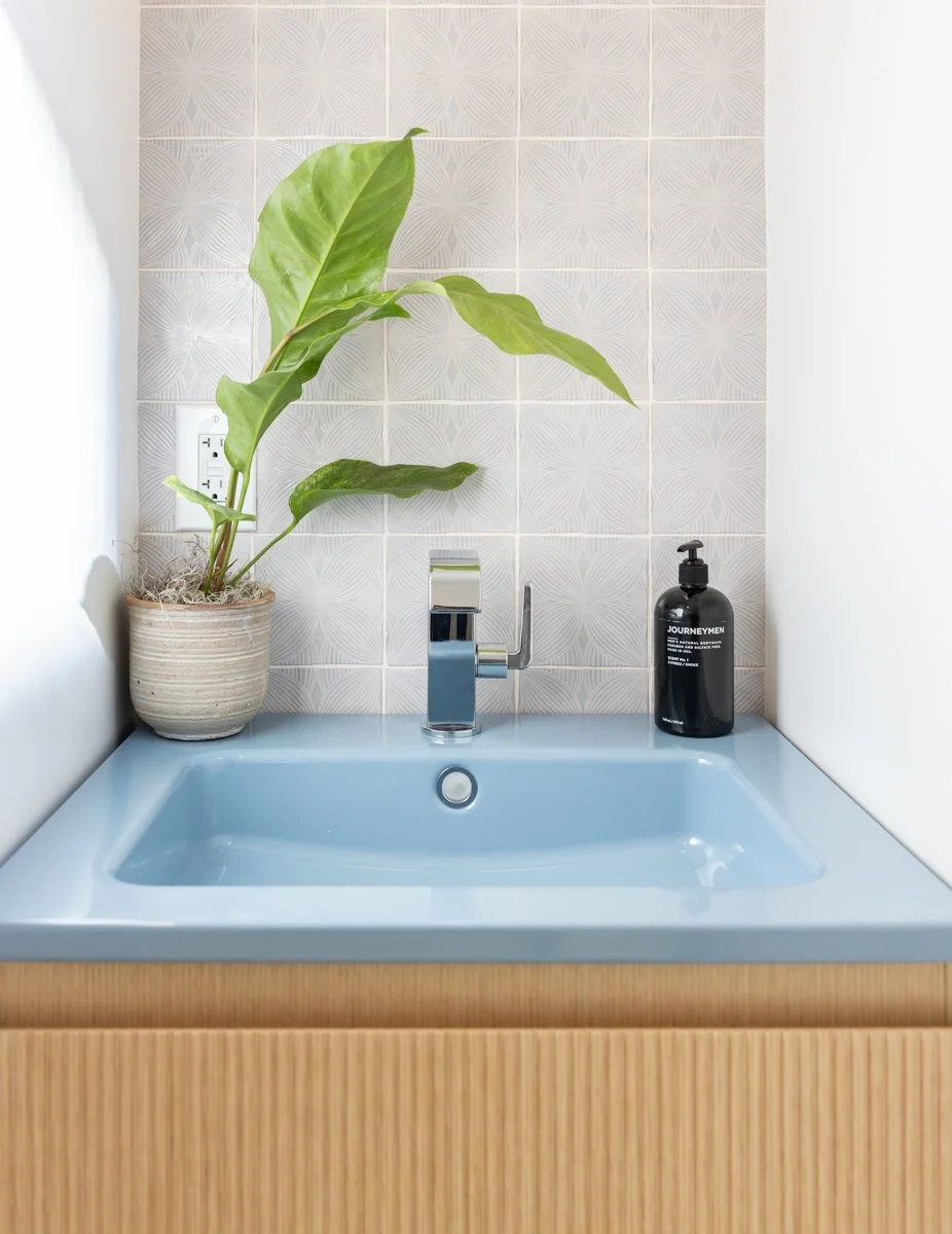A bathroom sink with a light blue basin, a modern chrome faucet, a potted plant with large green leaves, and a black soap dispenser on a wooden vanity, with textured white tiles in the background.