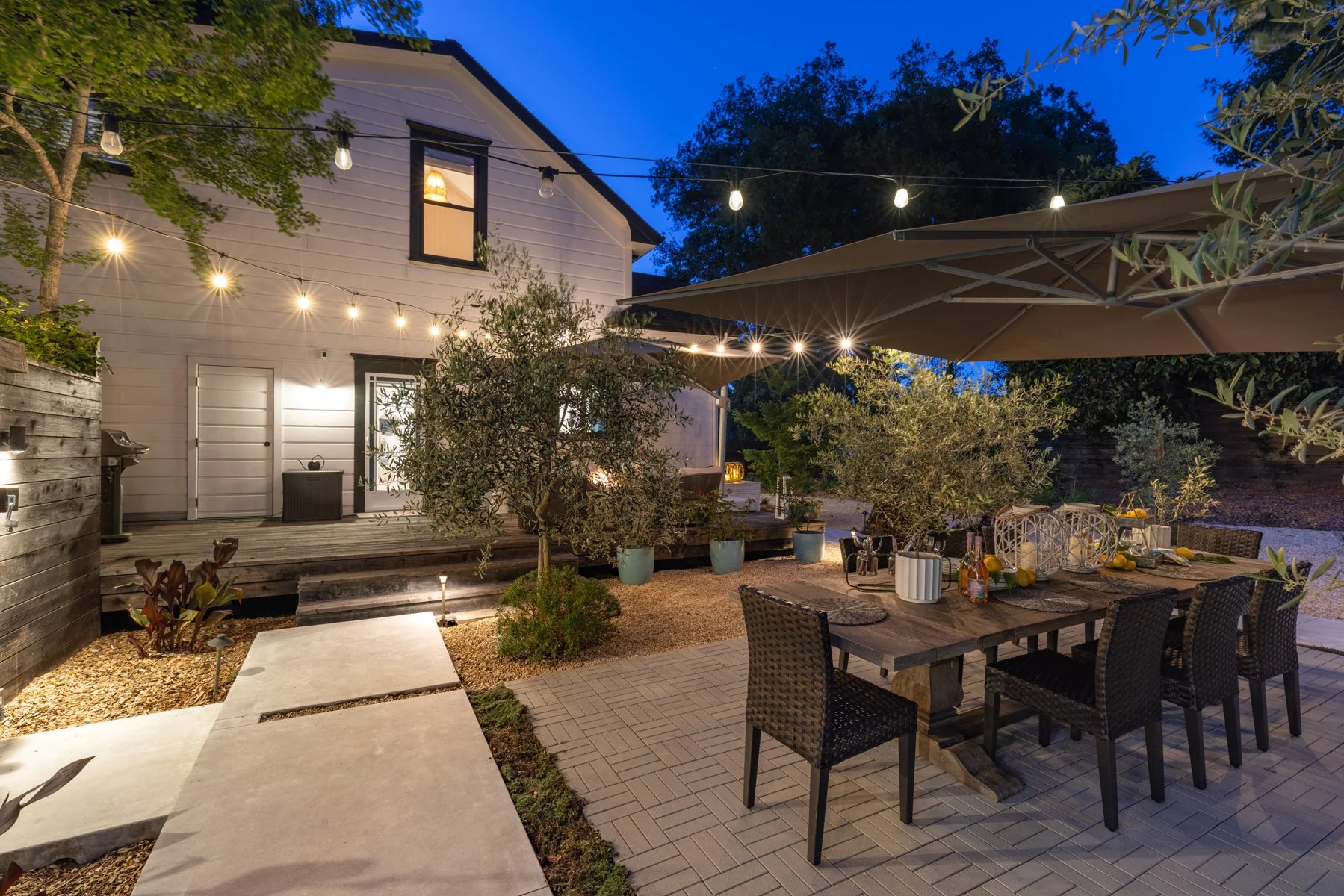 A cozy backyard patio illuminated by string lights at dusk, featuring a wooden table with chairs, potted plants, and an outdoor dining setup with lemons and candles.