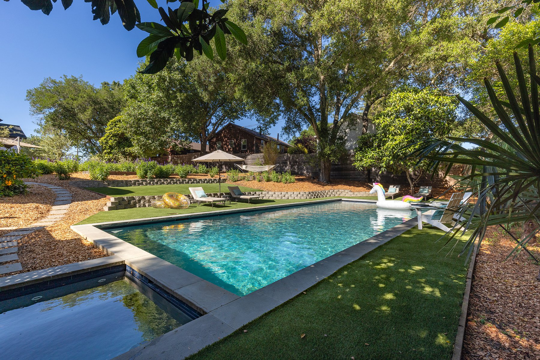 A backyard with a swimming pool surrounded by lounge chairs, a large umbrella, and lush green trees and plants under a clear blue sky.