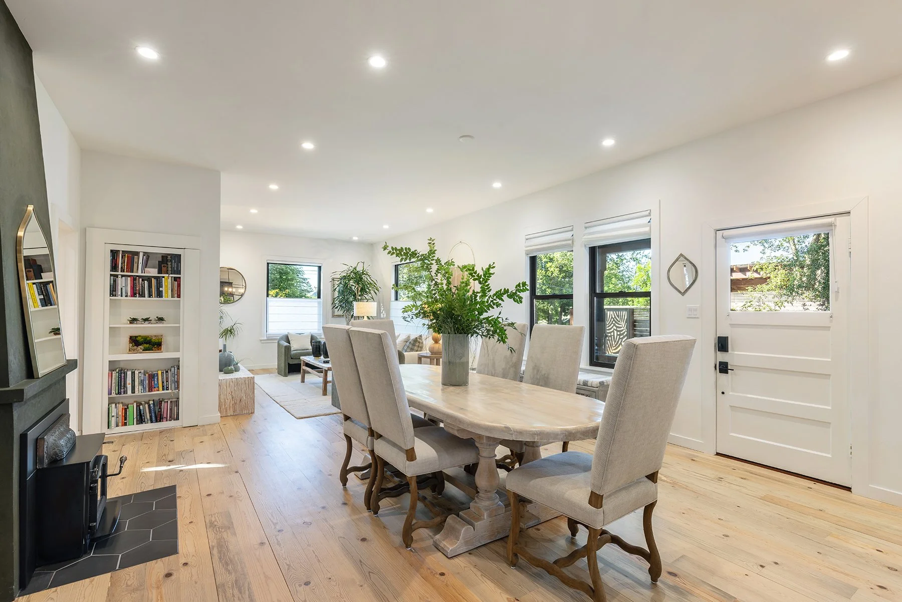 Bright, airy living and dining room with white walls, windows with blinds, a wooden dining table with upholstered chairs, a large green plant in the center, a bookshelf filled with books, and a sliding glass door leading outside.