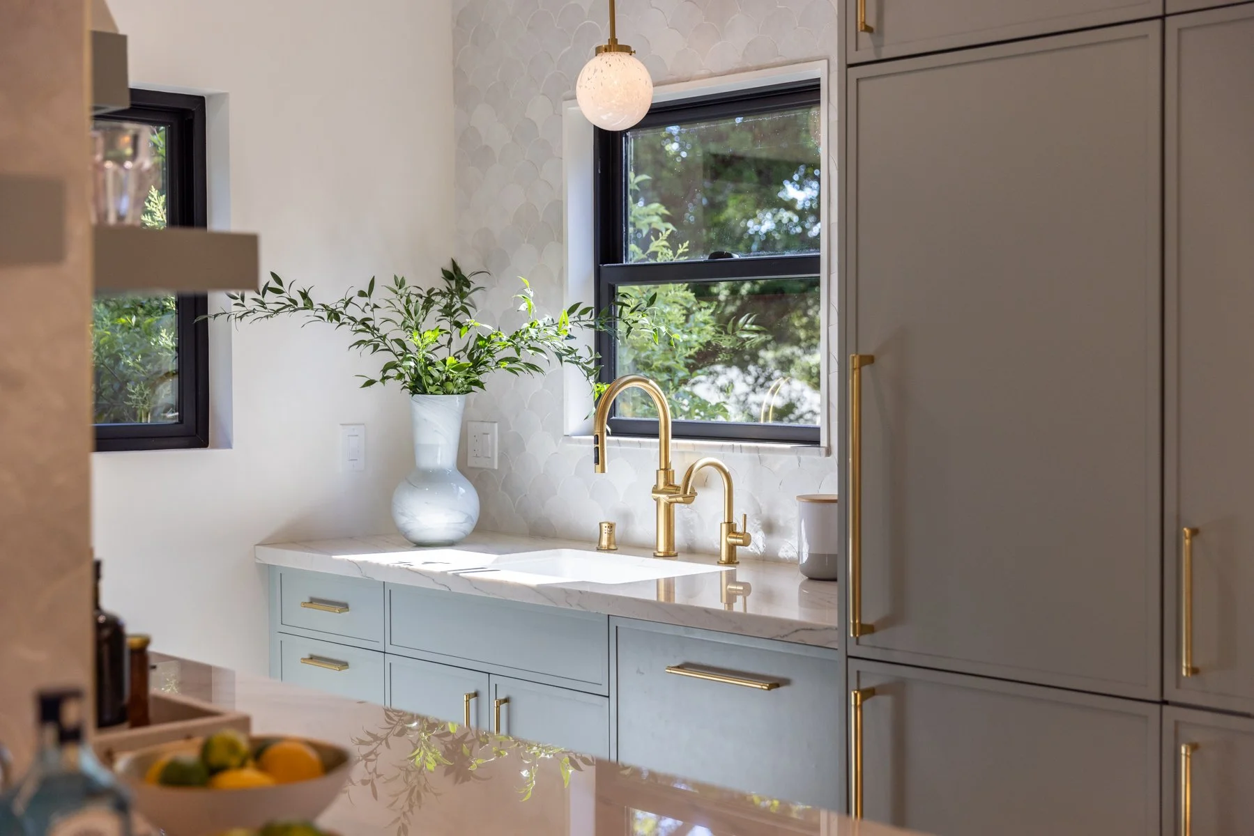 A modern kitchen with white marble countertops, blue cabinets with gold handles, gold faucet, and two large windows with black frames bringing in natural light. There is a white vase with green foliage on the counter and a pendant light hanging from 
