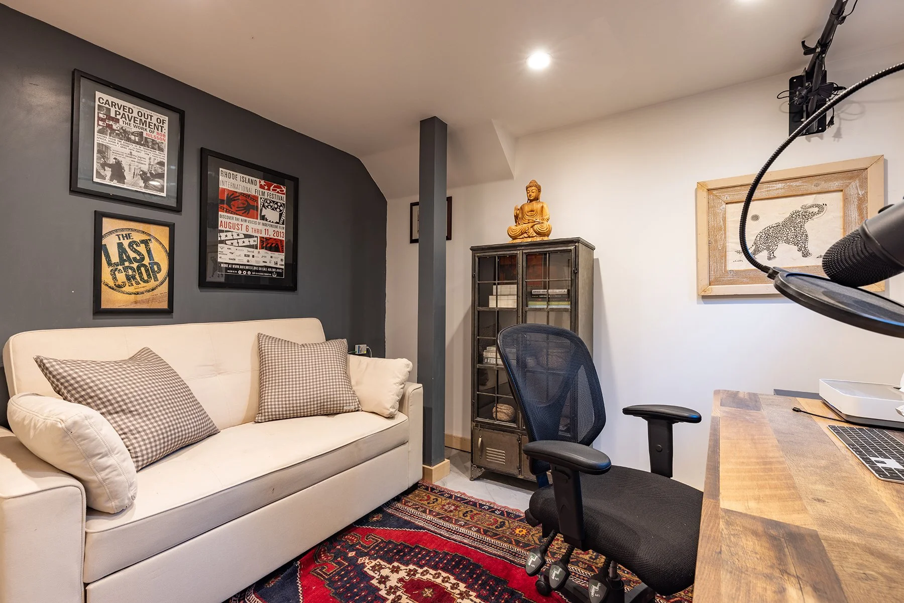 Interior of a cozy home office or studio space with a white sofa, a black office chair, and a wooden desk. Decor includes framed posters on the dark gray and white walls, a small glass cabinet with a Buddha statue on top, and a framed drawing of an e