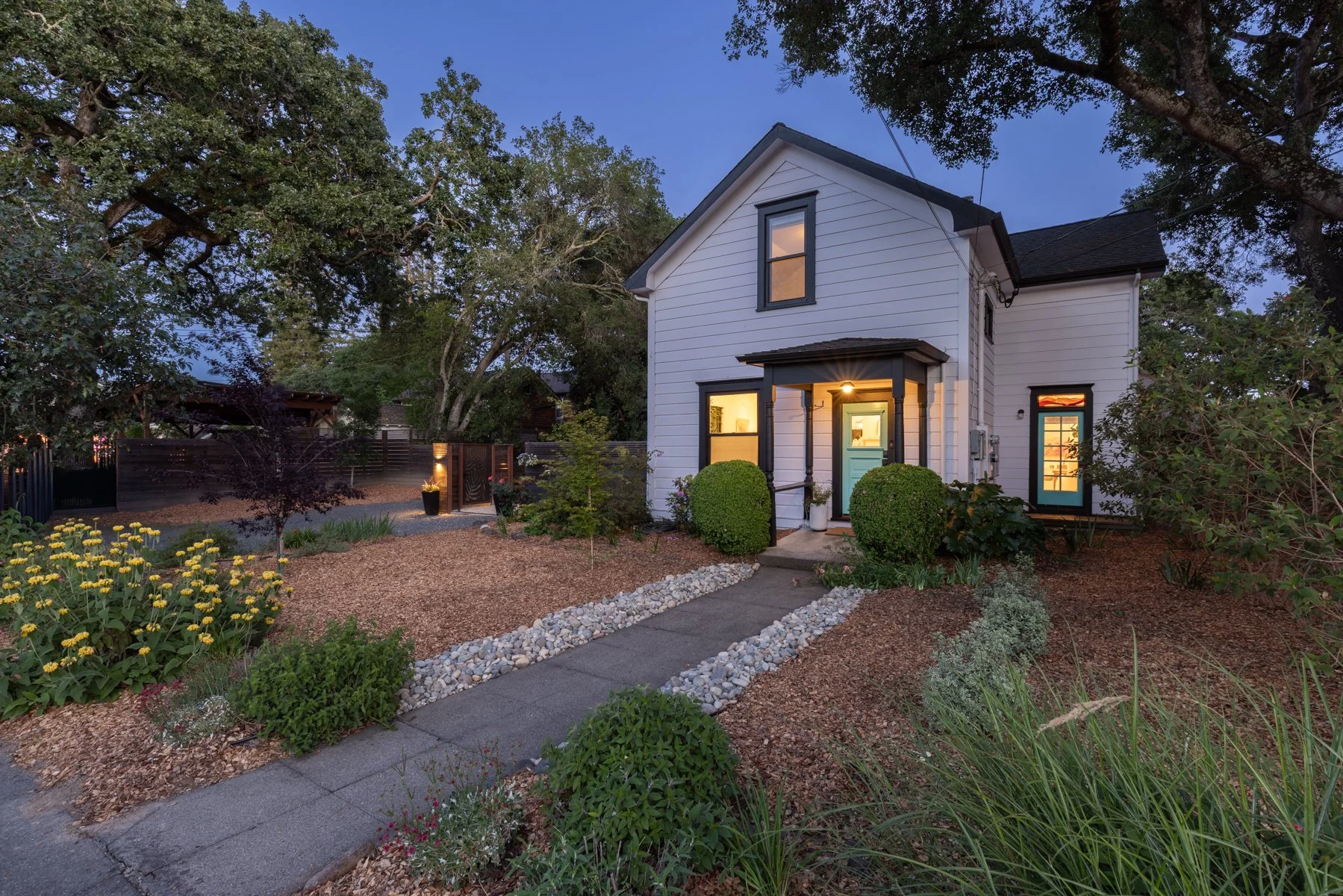 A modern two-story white house with black trim, showing a front porch with a light on, surrounded by a landscaped yard with bushes and flowers, trees, and a pathway with stone borders during evening or dusk.