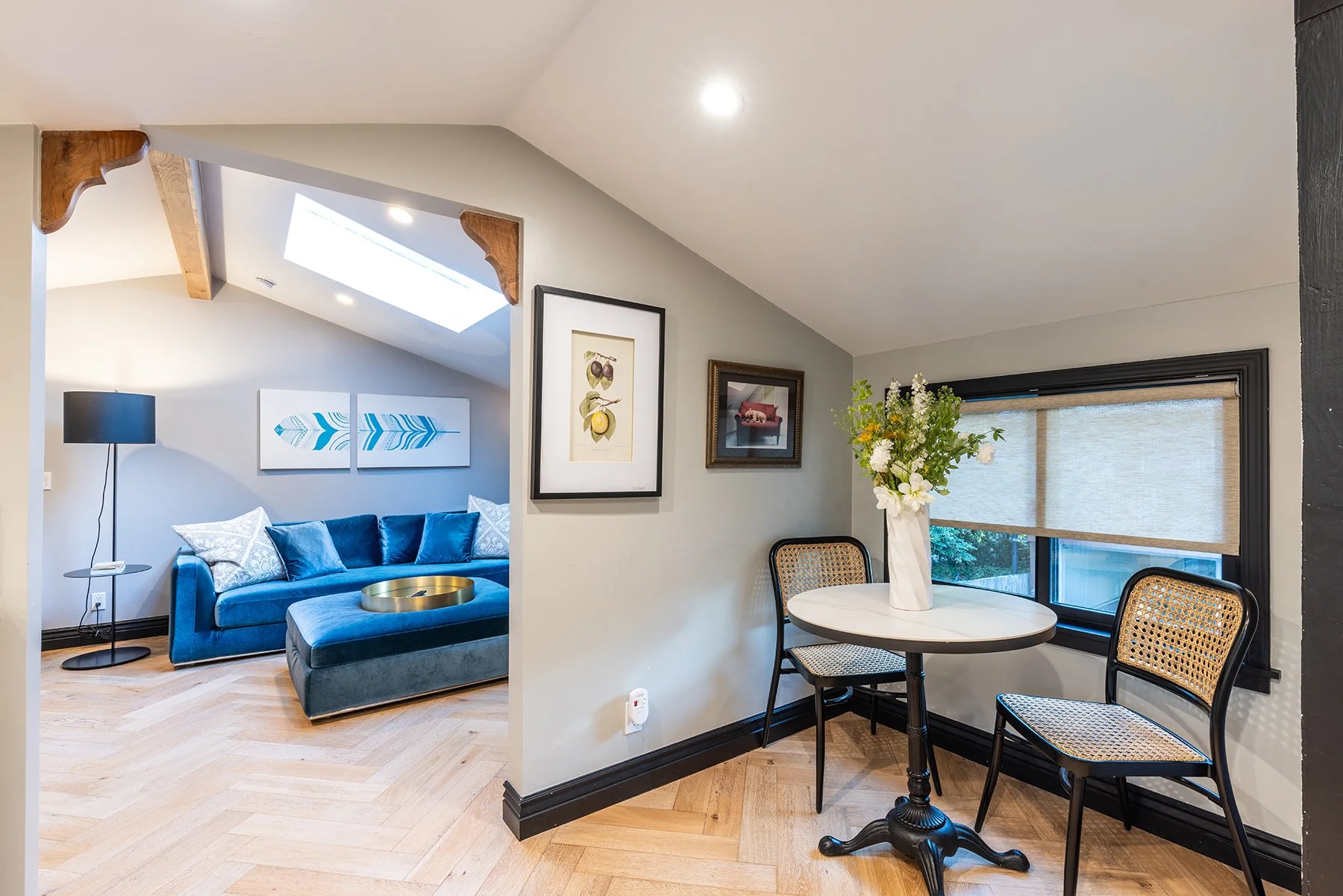 Living room with skylight, blue sofa, artwork on walls, and a dining area with a vase of flowers and two cane chairs.