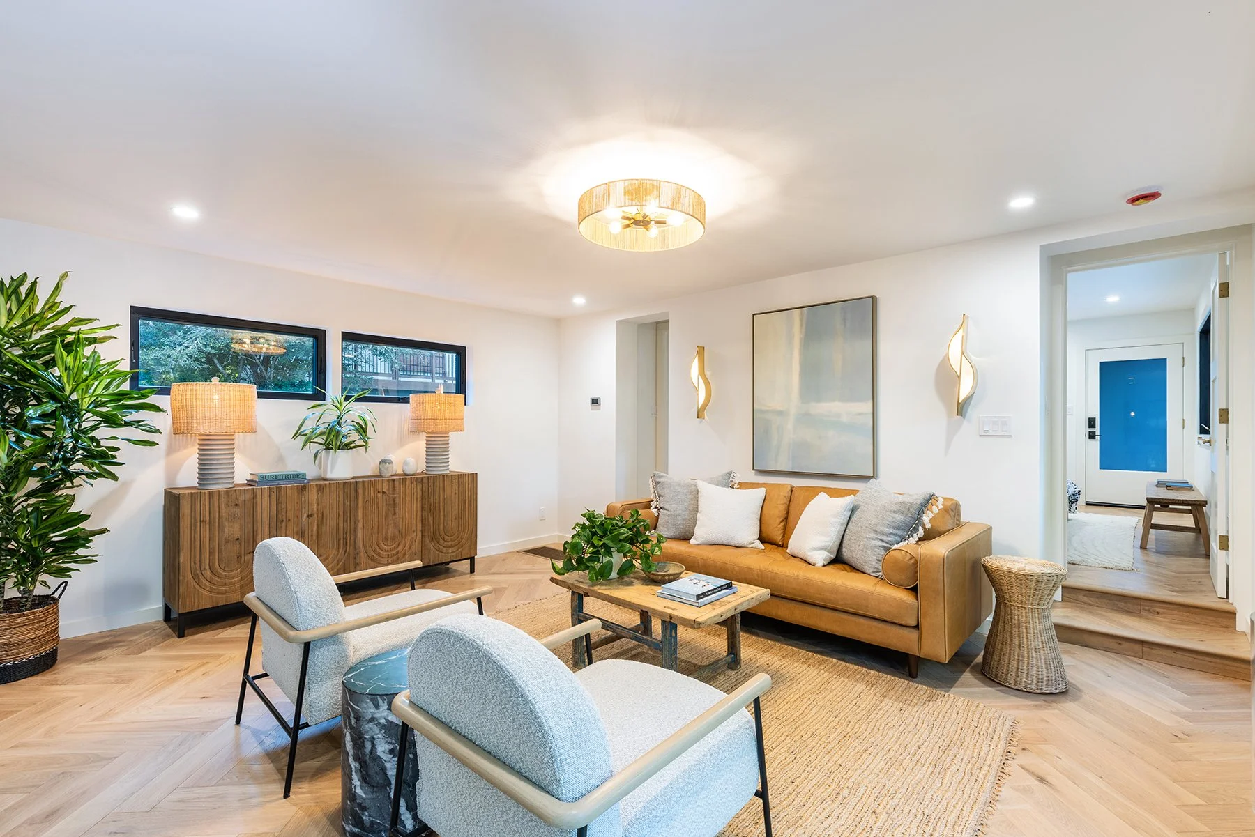 Living room with beige leather sofa, white armchairs, wooden coffee table, potted plants, artwork on the white walls, and modern light fixtures. Large windows and hardwood floors complete the cozy, stylish space.