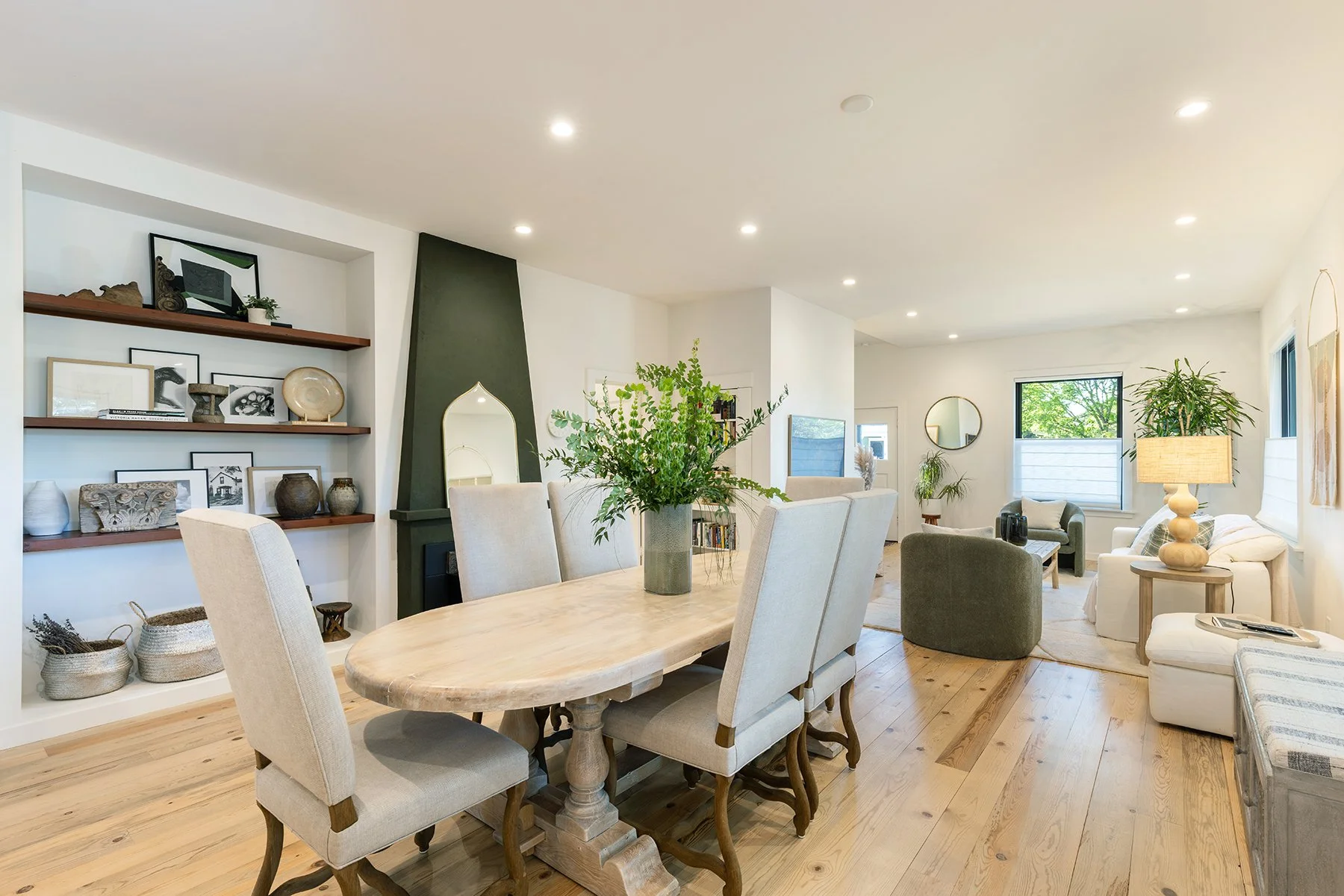 Open-concept living and dining room with wooden flooring, a dining table with beige upholstered chairs, a large green plant in the center, white and gray sofas, side tables with lamps, windows with shades, and wall-mounted decor.