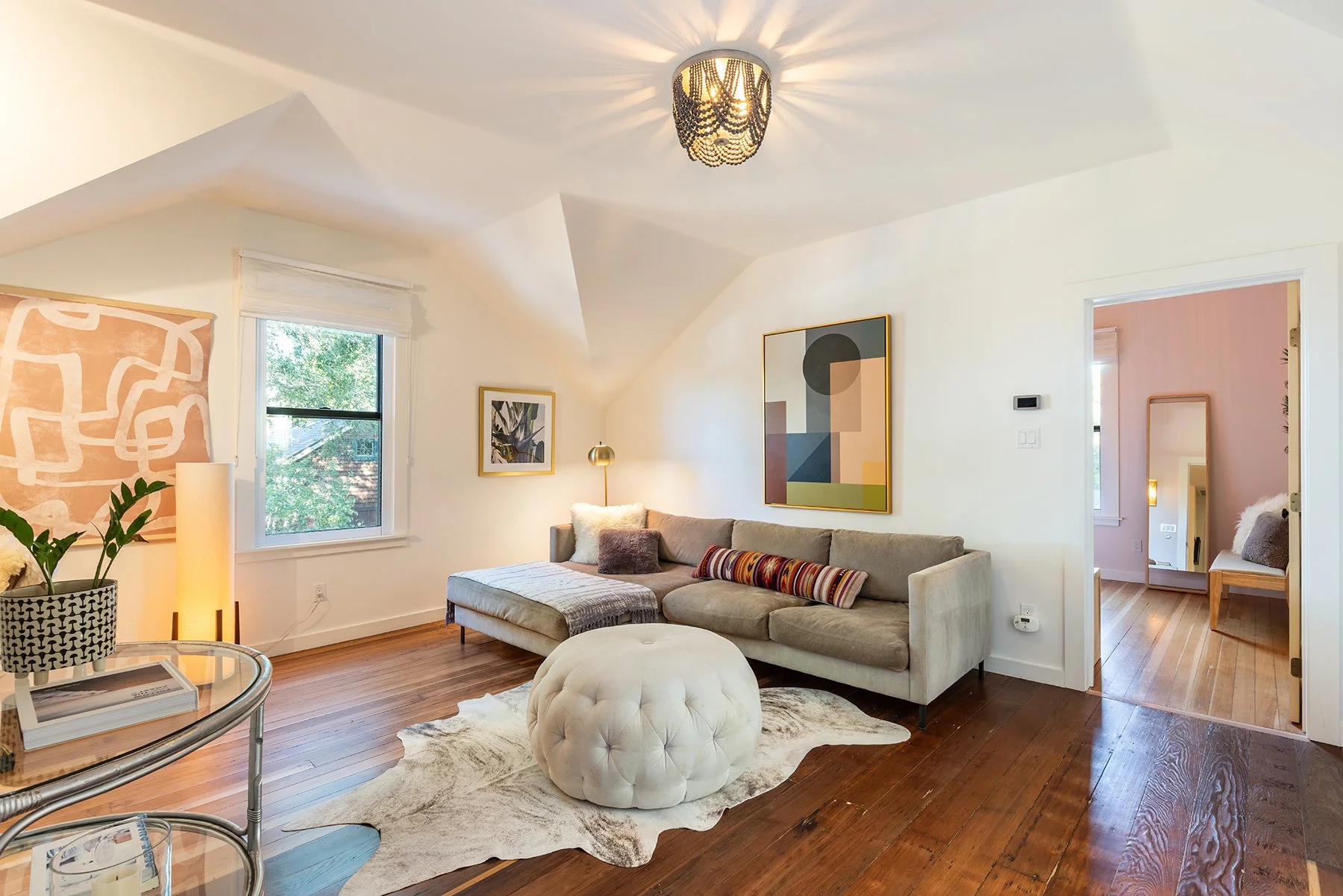 Living room with beige sectional sofa, colorful pillows, and a round tufted ottoman on a cowhide rug. Wall art, a gold lamp, a decorative plant, and a window with a view of trees are visible.