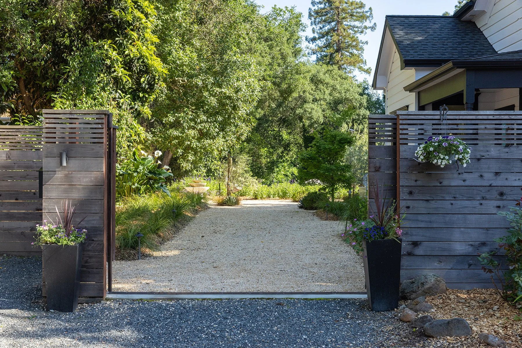 A gravel driveway leading through a wide wooden gate into a landscaped garden with lush green trees, potted plants, and a white house with dark roof and siding in the background.