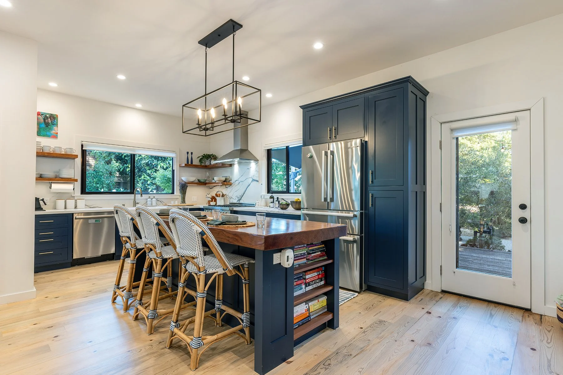 Modern kitchen with blue cabinets, wooden island, white walls, hardwood floors, open shelving, stainless steel fridge, window and glass door leading outside, black chandelier with candle-like lights.