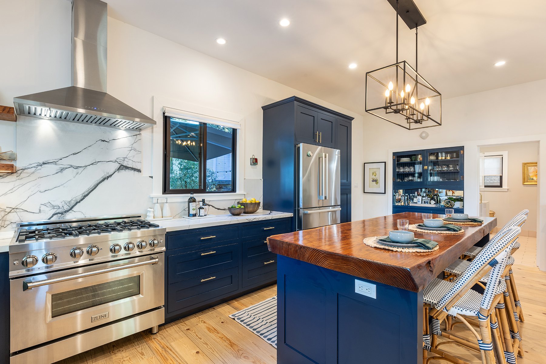 A modern kitchen with navy blue cabinets, a wooden island, stainless steel appliances, a marble backsplash, a window, hanging light fixture, and dining chairs with patterned cushions.