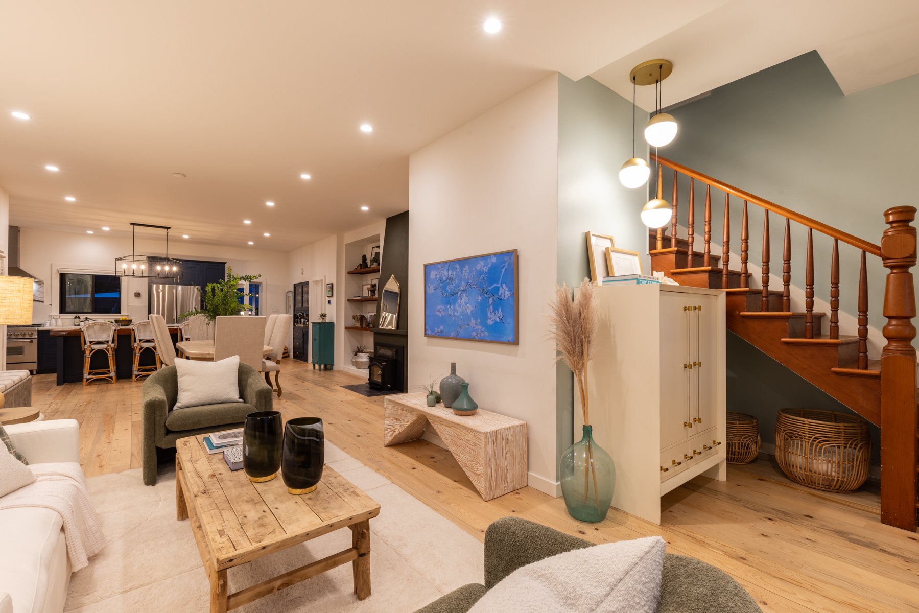 Open-concept living area with kitchen, dining, and living space featuring wooden floors, beige furniture, and modern decor. A staircase with wooden railing is visible on the right, with lighting fixtures hanging from the ceiling.