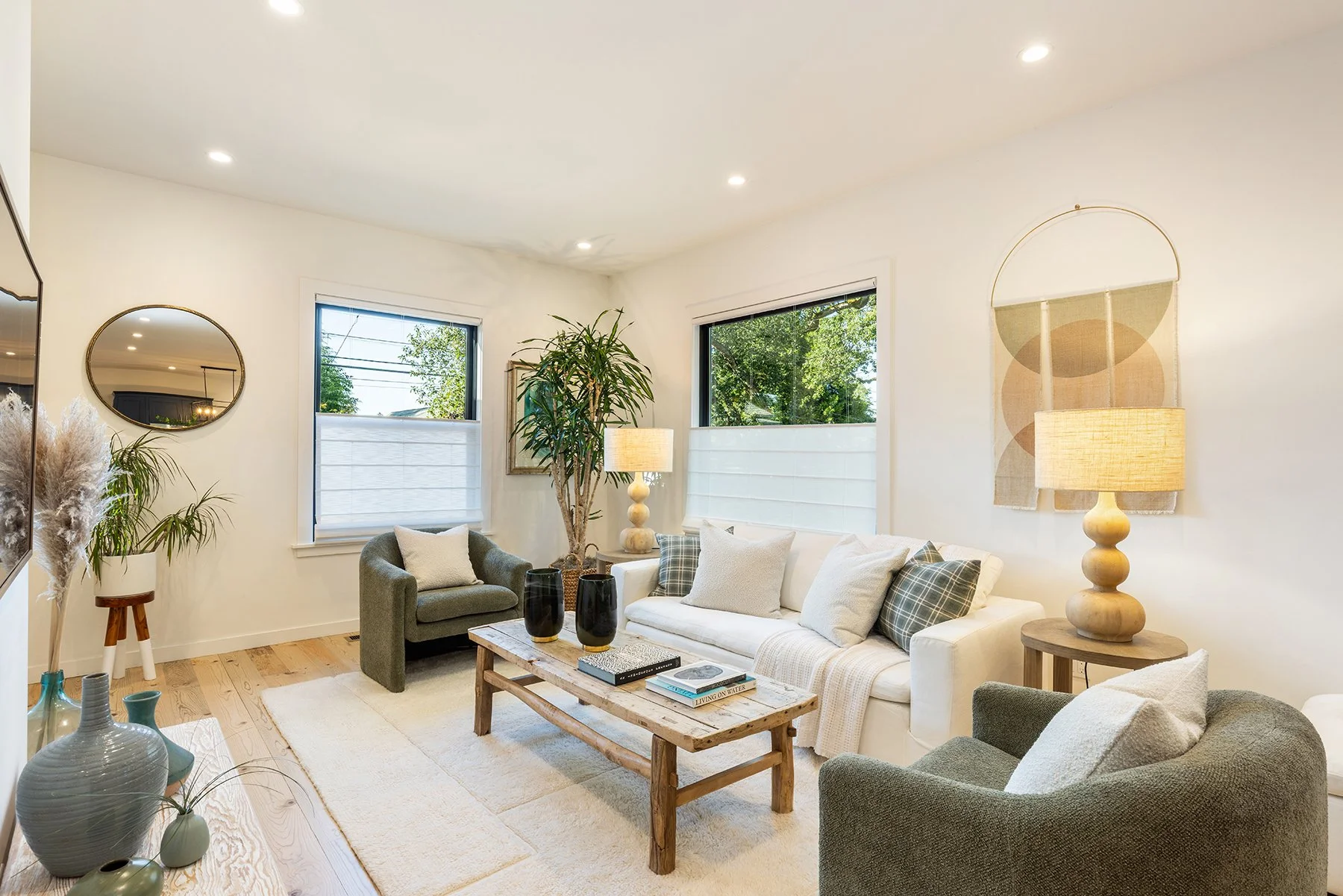 Living room with white walls, beige carpet, and natural light from multiple windows. Contains a white sofa with pillows, a green armchair, a wooden coffee table with books and black vases, various lamps, and indoor plants. Decorative wall art and lar