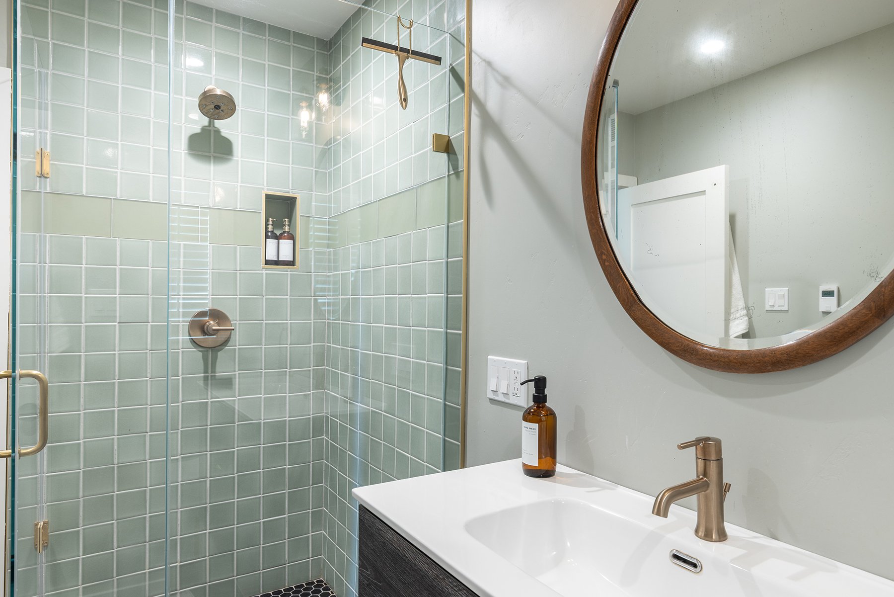 Modern bathroom with a glass-enclosed shower featuring green tiles, a built-in niche with soap dispensers, and a white vanity with a brass faucet. There is a large round mirror on the wall, a soap pump on the vanity, and a wall-mounted towel.