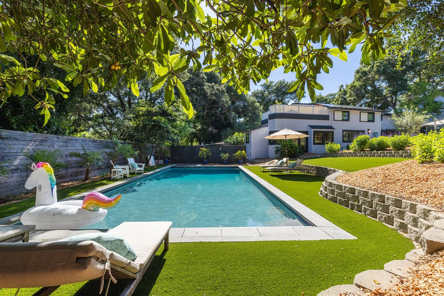 A backyard with a swimming pool, pool float shaped like a unicorn, lounge chairs, an umbrella, and a modern house in the background. Tree branches with green leaves hang overhead.