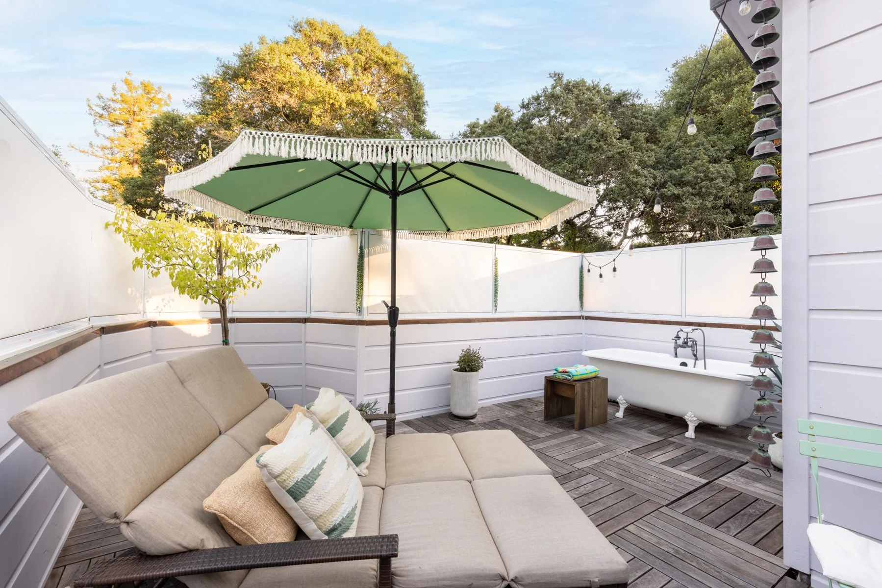 An outdoor patio with beige sectional sofa, decorative pillows, a large green patio umbrella with white fringe, a small potted tree, and a vintage clawfoot bathtub in the corner, surrounded by white fencing and string lights, with trees in the backgr