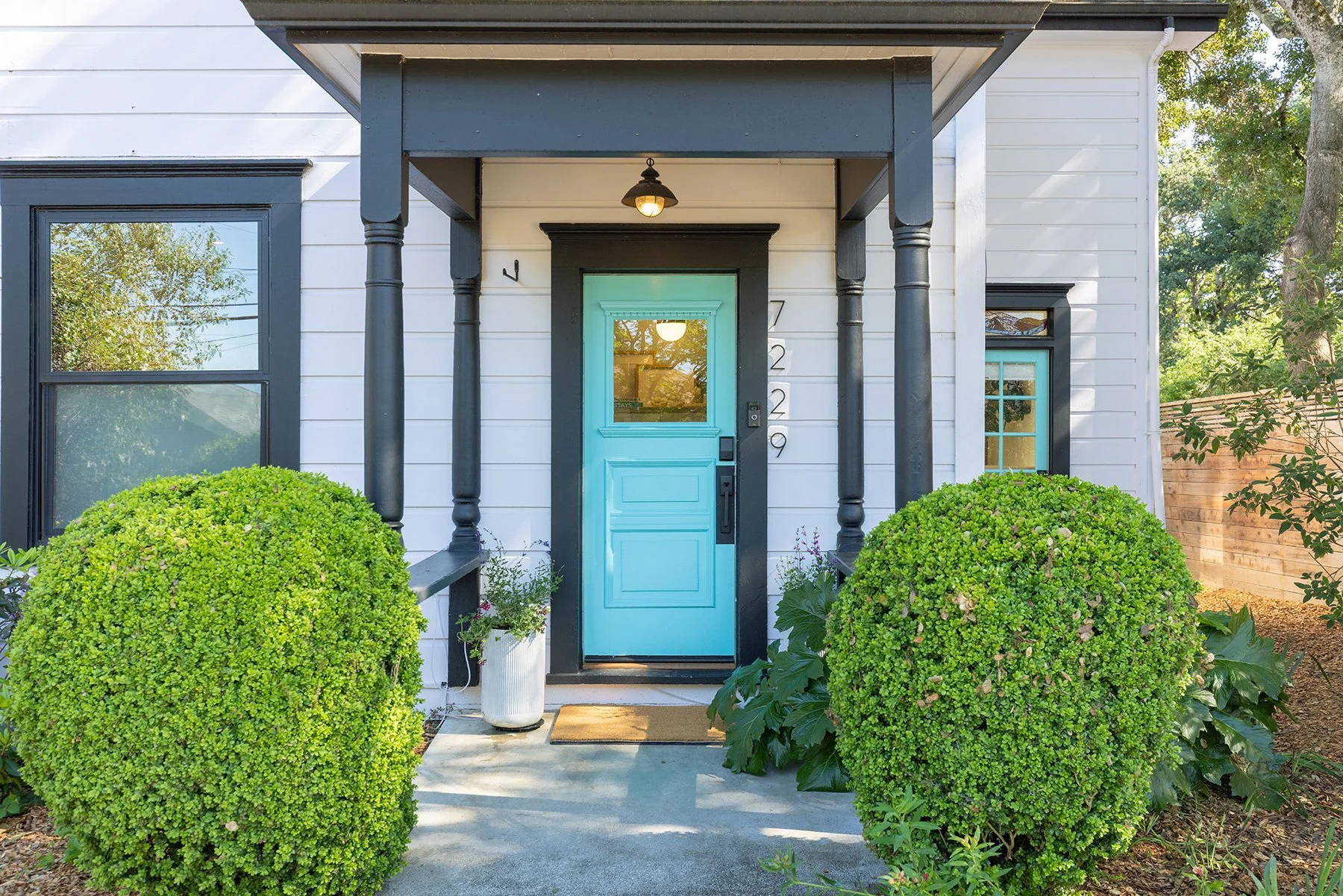 Front entrance of a house with a light blue door, black trim, and a small porch supported by black columns. Two large, rounded green bushes are on each side of the walkway leading to the door, with potted plants beside the steps. The house exterior i