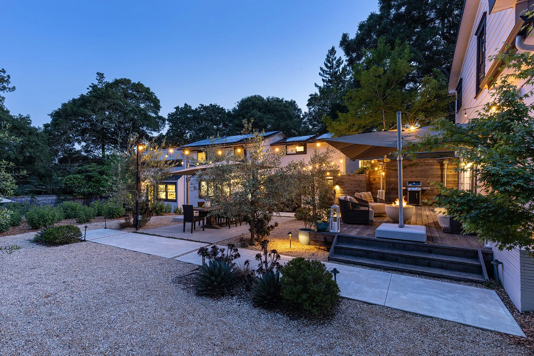 A backyard at dusk with a wooden deck, outdoor furniture, string lights, and a fire pit. There are trees and a house with windows and a large umbrella.