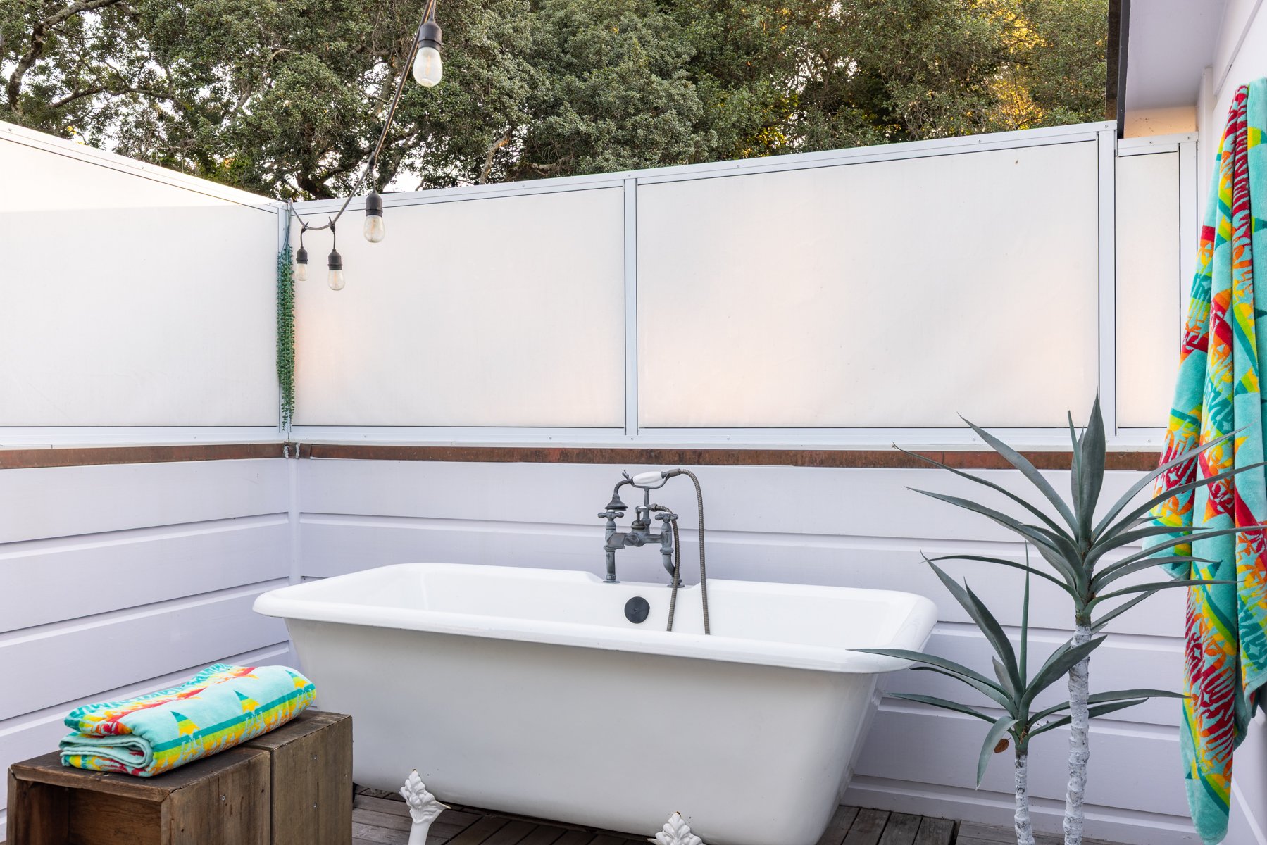 Outdoor bathtub area with a white clawfoot bathtub, a wooden side table with colorful towels, colorful towels hanging on a wall, a potted plant, and string lights, with trees in the background.