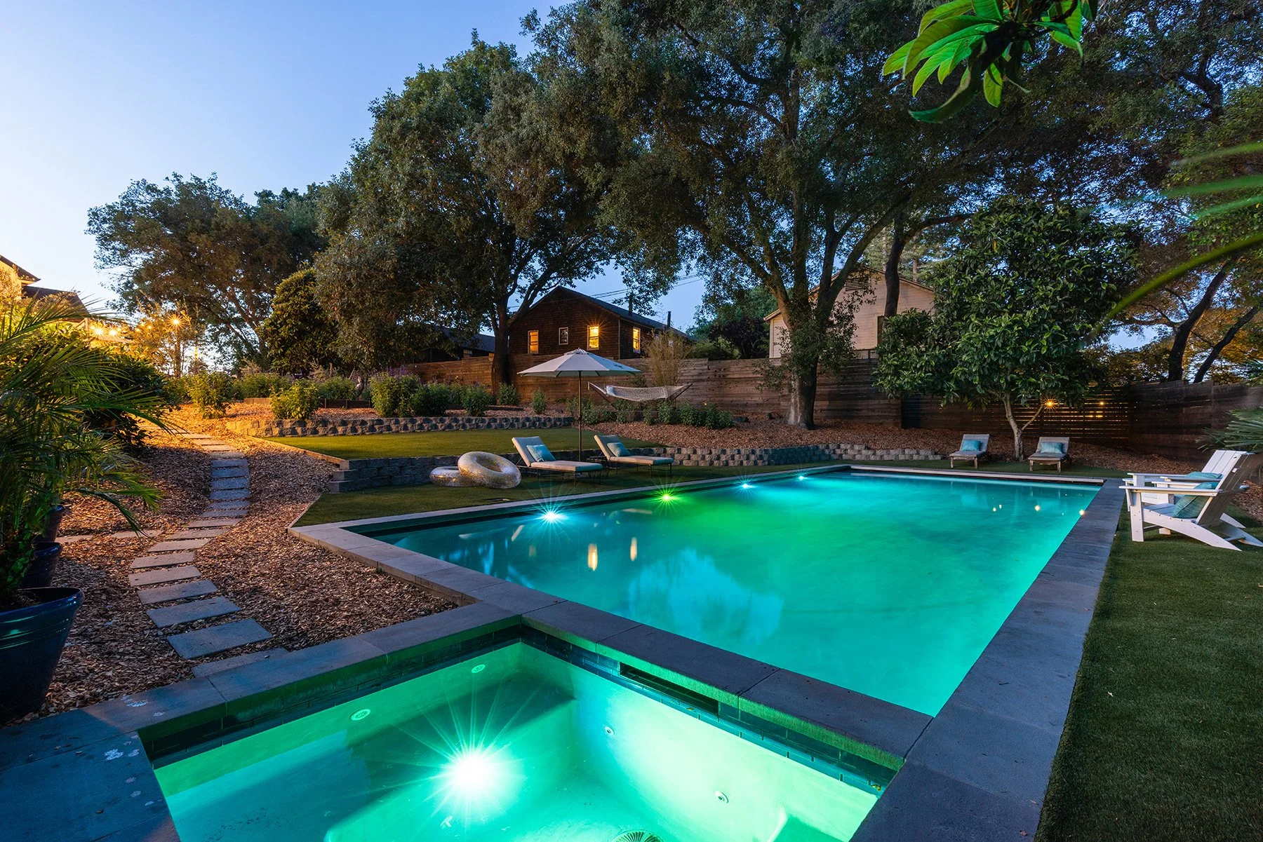 A backyard at dusk with a lit swimming pool surrounded by lounge chairs, trees, and a wooden fence, with a house in the background and outdoor lighting.