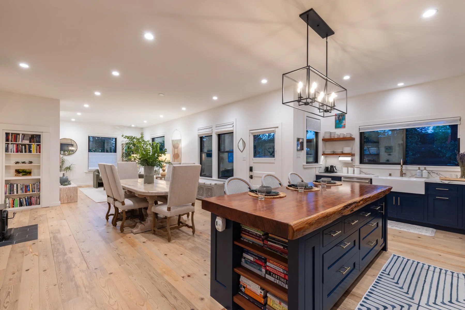 Spacious open-concept kitchen and dining area with wooden flooring, white walls, and large windows. The kitchen features a large wooden island with a bookshelf underneath, black cabinets, and open shelving with white dishes. A modern chandelier hangs