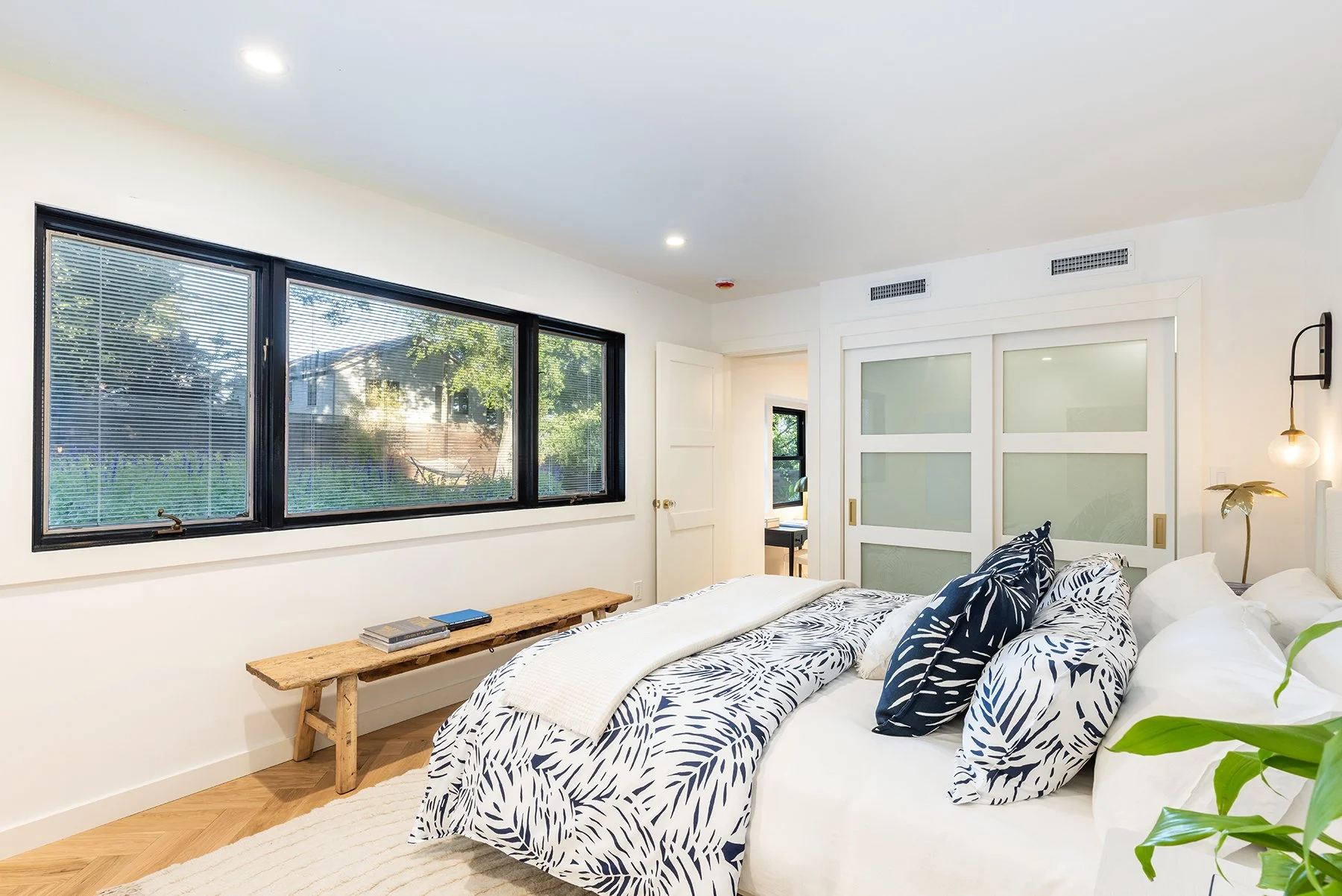 Bedroom with large windows, a bed with black and white leaf-patterned bedding, decorative pillows, a wooden bench, and greenery outside.
