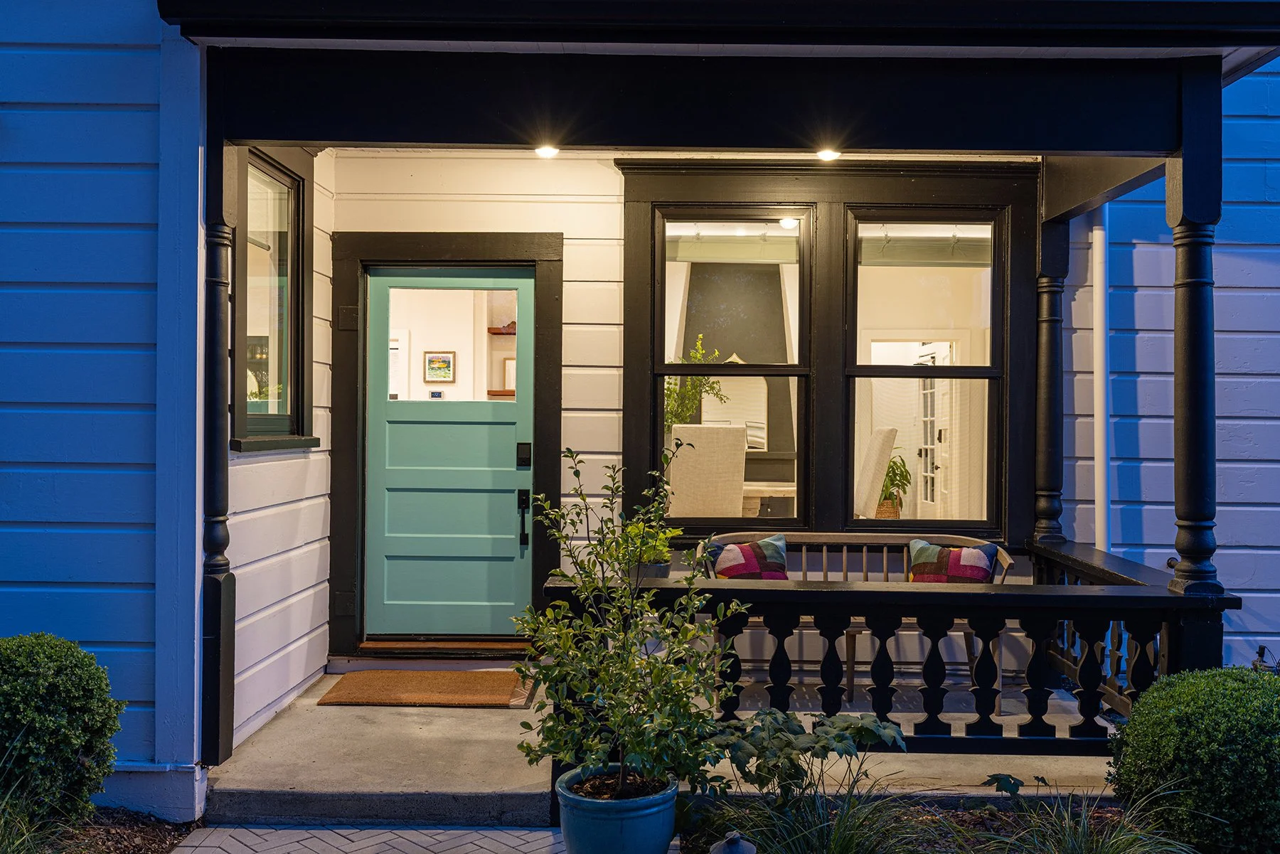 Front porch of a house with a green door, black trim, and a small porch with a wooden bench and potted plants, illuminated by interior and overhead lights at night.