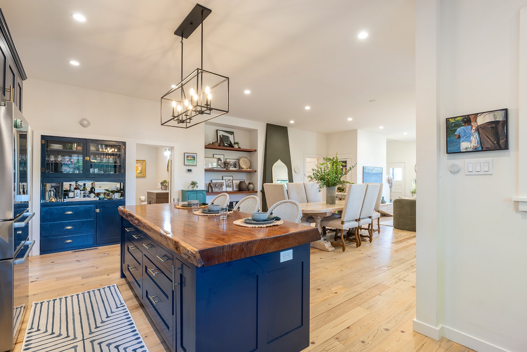 Open-concept kitchen and dining area with blue cabinets, a large wooden island, beige chairs around a dining table, and decorative shelving; modern lighting and wooden flooring.