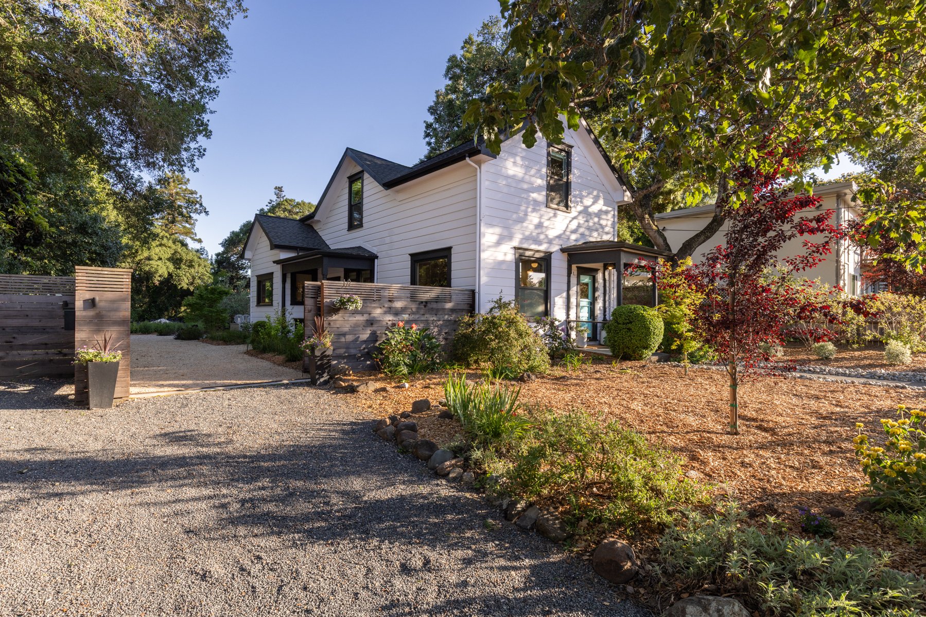 A modern white two-story house with black window frames and a black roof, surrounded by a well-maintained garden with various plants, flowers, and trees. There is a gravel driveway leading up to the house, and a wooden fence with flower pots.