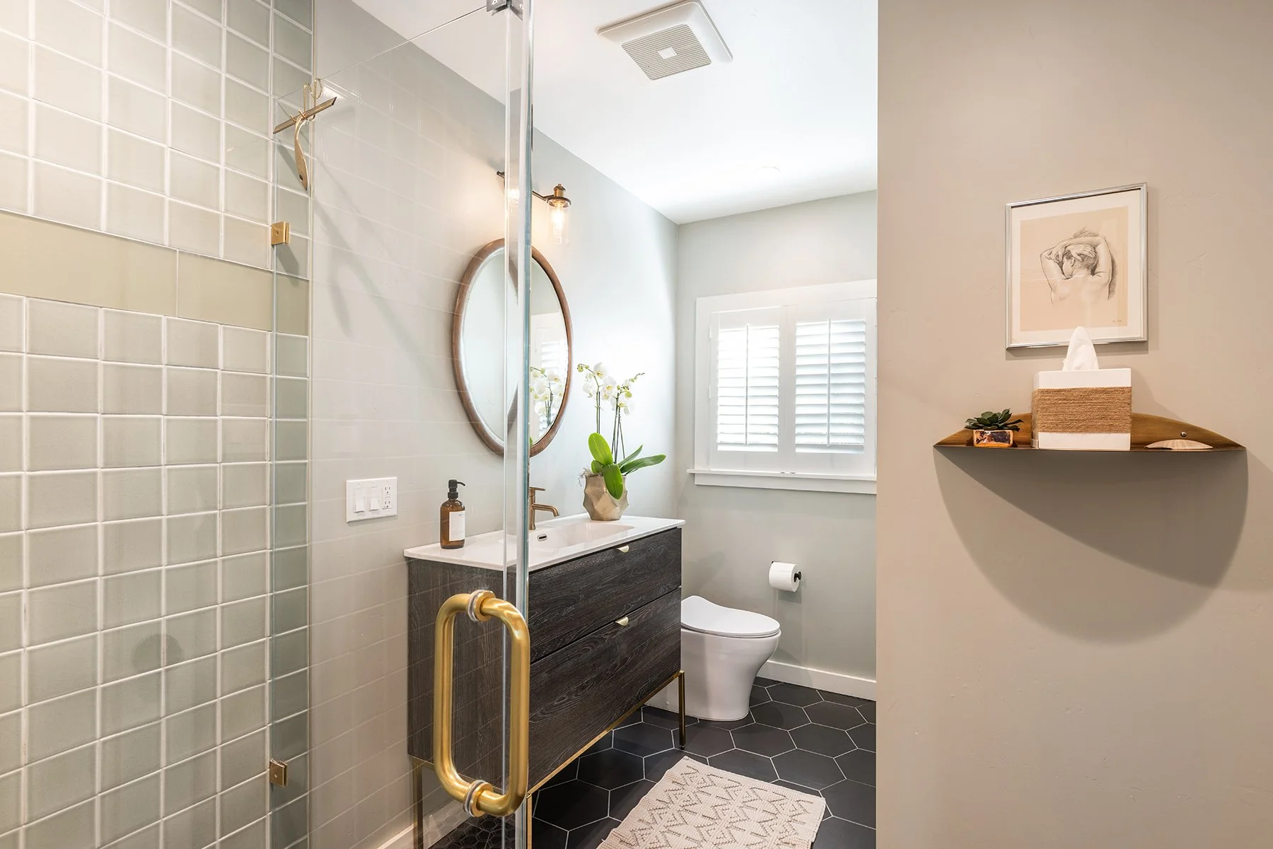 Modern bathroom with black hexagonal floor tiles, a white toilet, a dark wooden vanity with a white countertop and an oval mirror, a potted orchid plant, wall-mounted light fixtures, a framed art print, a floating shelf with tissues and a plant, and 
