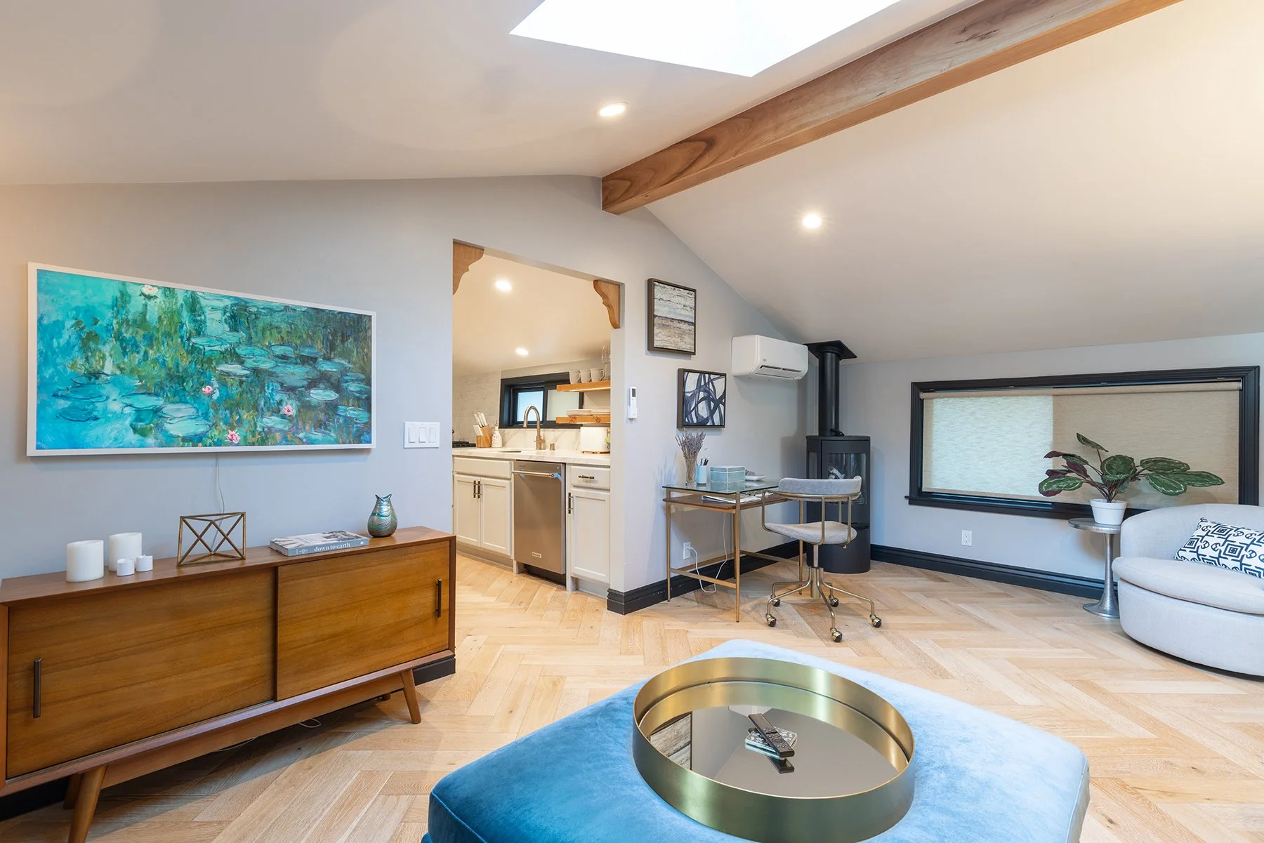 Modern living room with a blue velvet ottoman in the foreground with a gold tray and remote control, a wooden sideboard with candles and decor, a large abstract painting on the wall, a small kitchen area with beige cabinets, and an office corner with