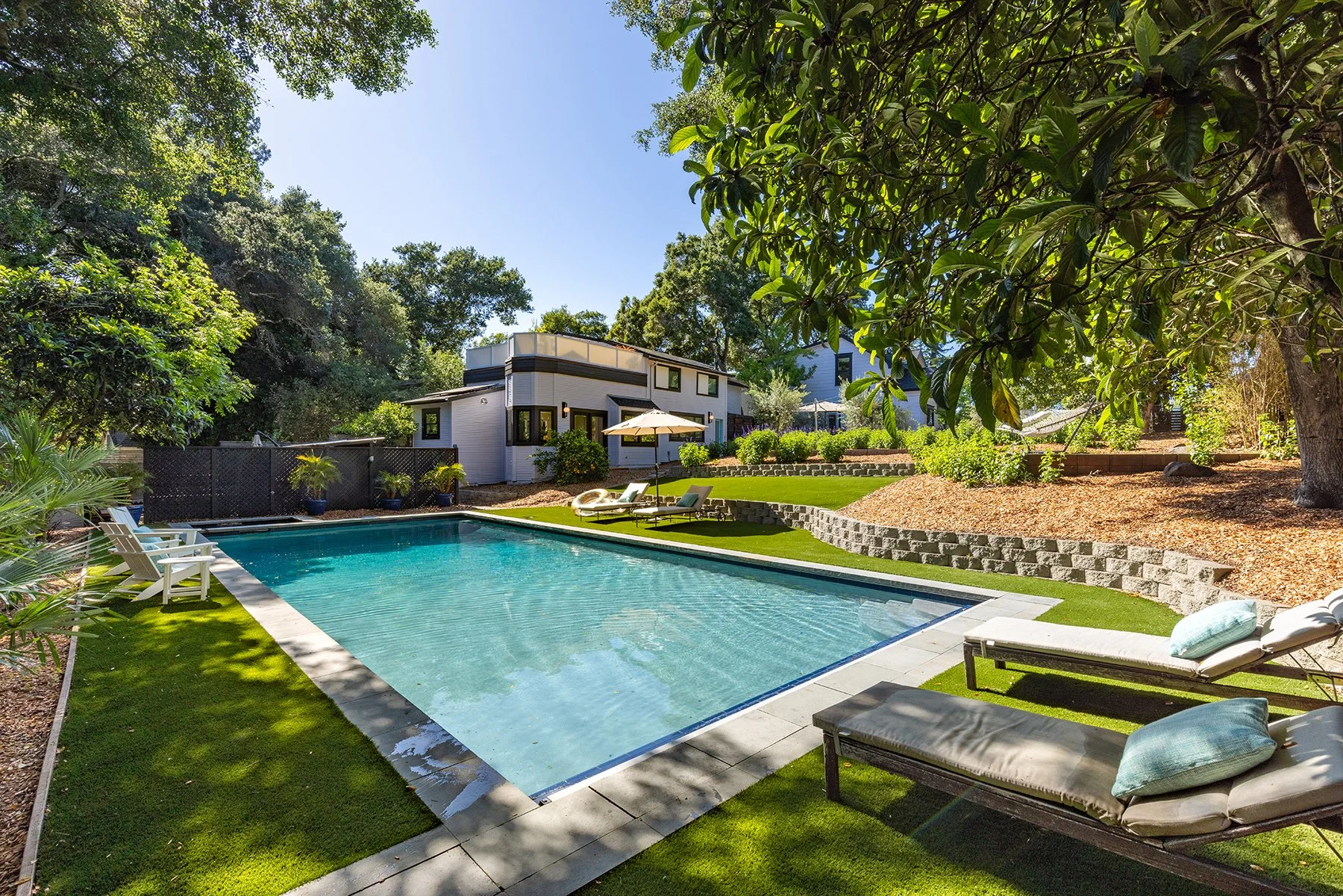 A backyard with a swimming pool, surrounded by green grass, lounge chairs, and umbrellas. There are trees and a modern house in the background, with a clear blue sky overhead.