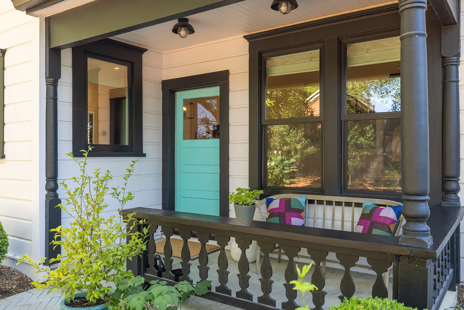 Front porch of a house with black trim and a mint green front door, black-paned windows, a wooden bench with colorful cushions, potted plants, and decorative elements.
