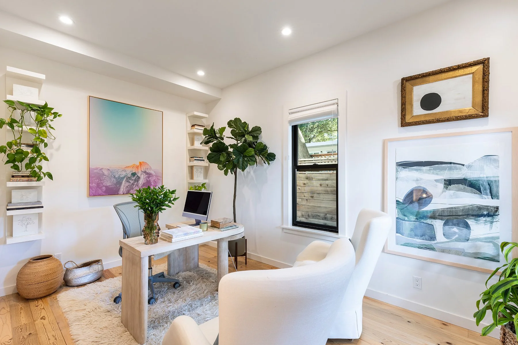 Bright modern home office with white walls, large windows, artwork, houseplants, and a wooden desk with a computer and books.