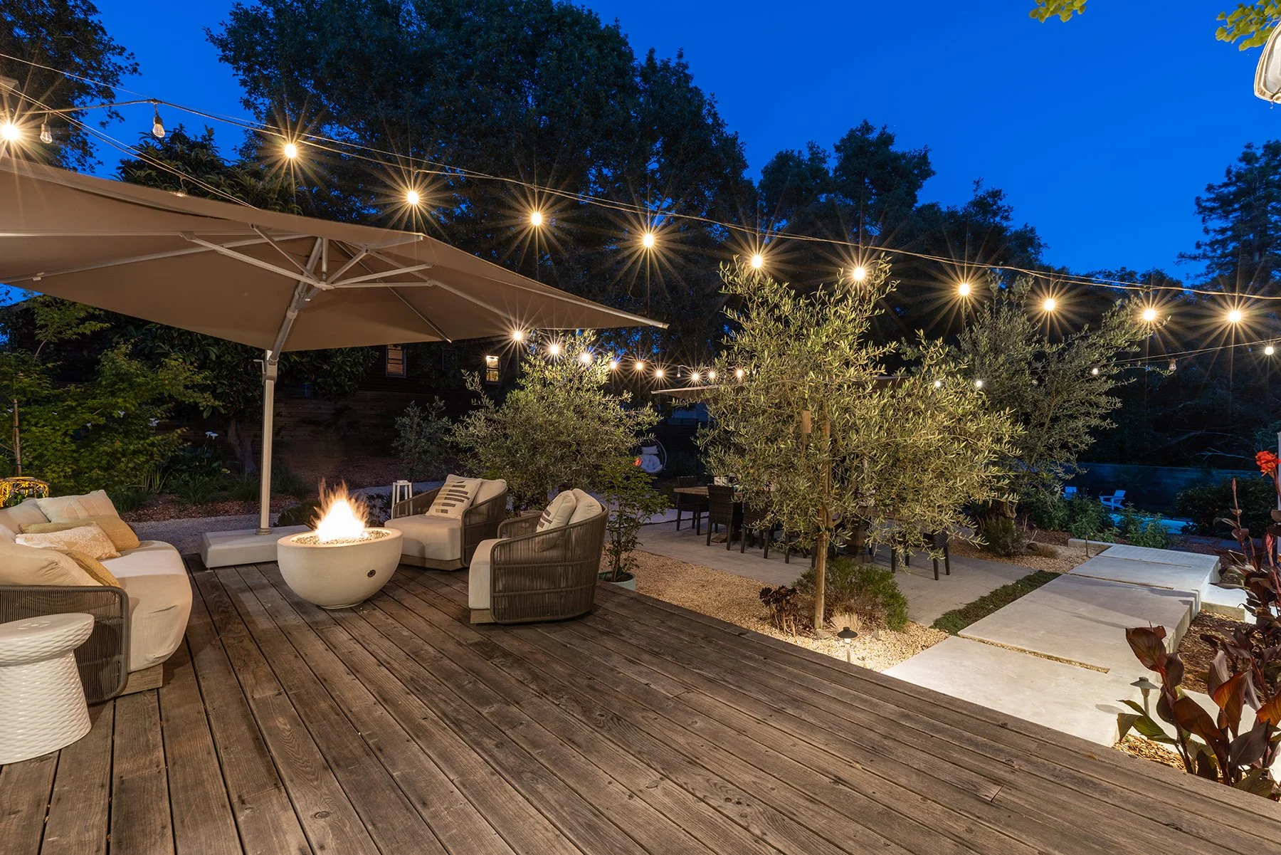 Outdoor patio at dusk with string lights overhead, a fire pit, outdoor seating with cushions, a large umbrella, trees, and a backyard with a swimming pool in the background.