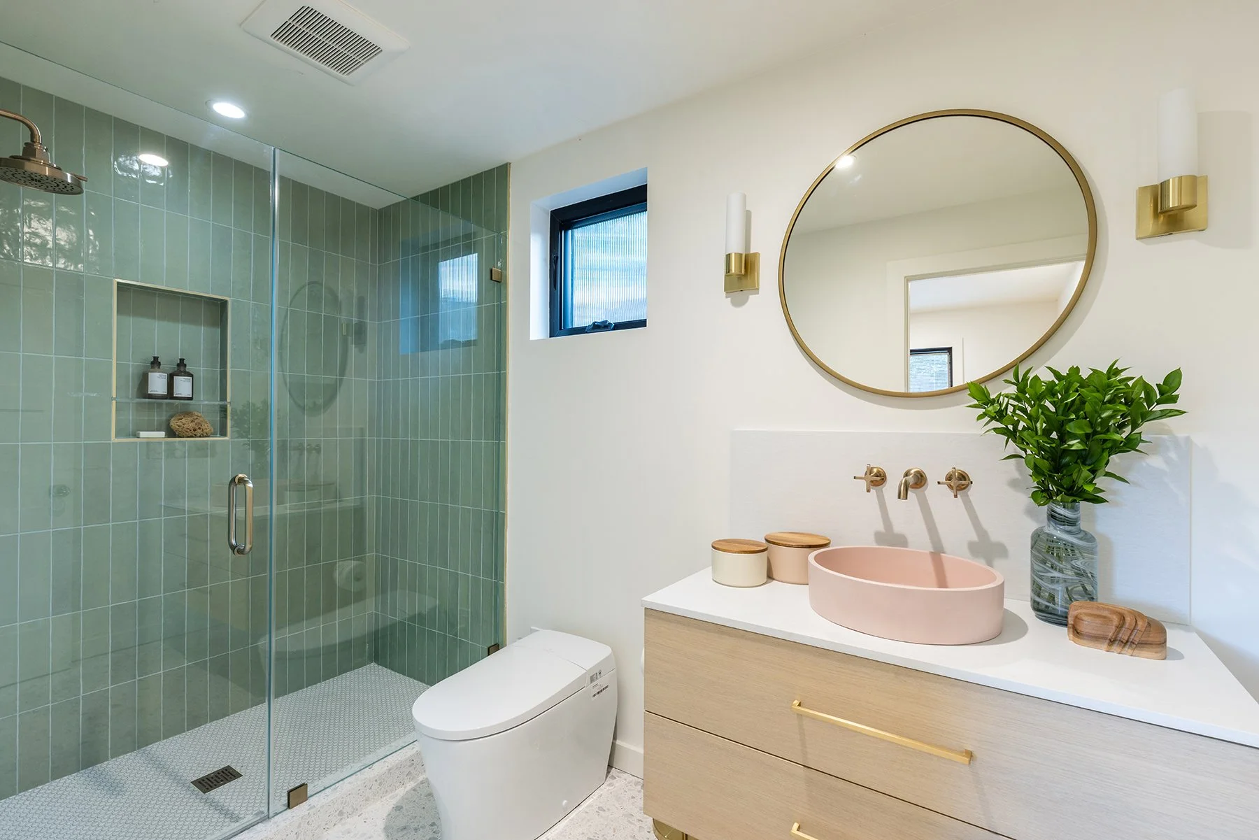 Modern bathroom with a glass-enclosed shower with green tiles, a round pink sink, a wooden vanity with gold hardware, a large round mirror, a potted green plant, a window, and a white toilet.