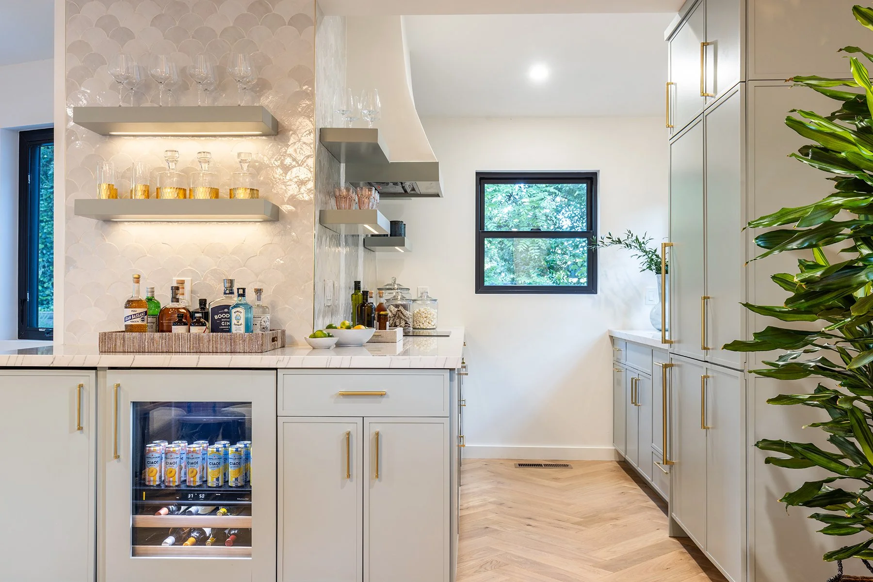 Modern kitchen with white cabinets with gold handles, a window overlooking greenery, open shelves holding glassware, a small beverage fridge, and a counter with bottles and bowls of fruit, with large green plant on the right.