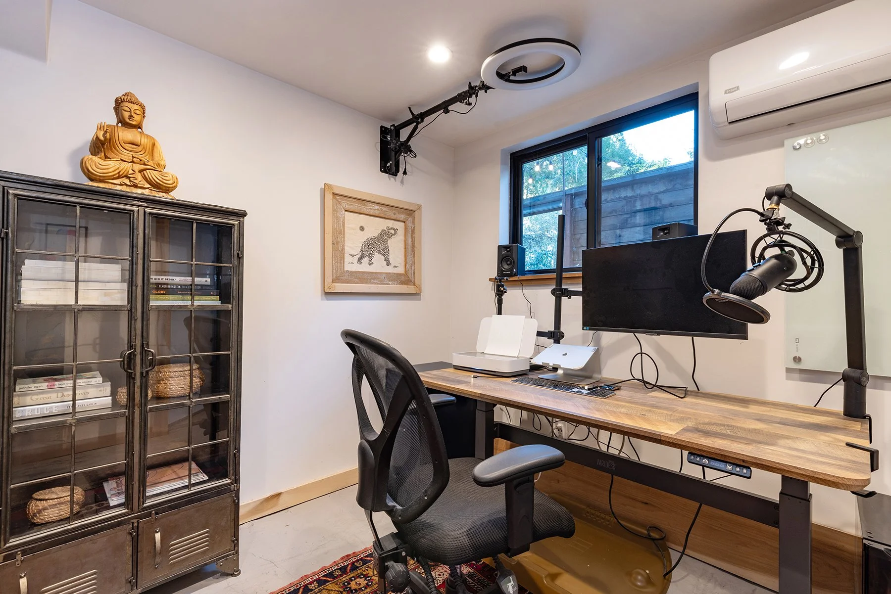 Home office with a wooden desk, black office chair, computer monitor, microphone on adjustable arm, and wall-mounted air conditioner. A window with a view outside, framed artwork of an elephant, and a Buddha statue on a vintage cabinet are also visib