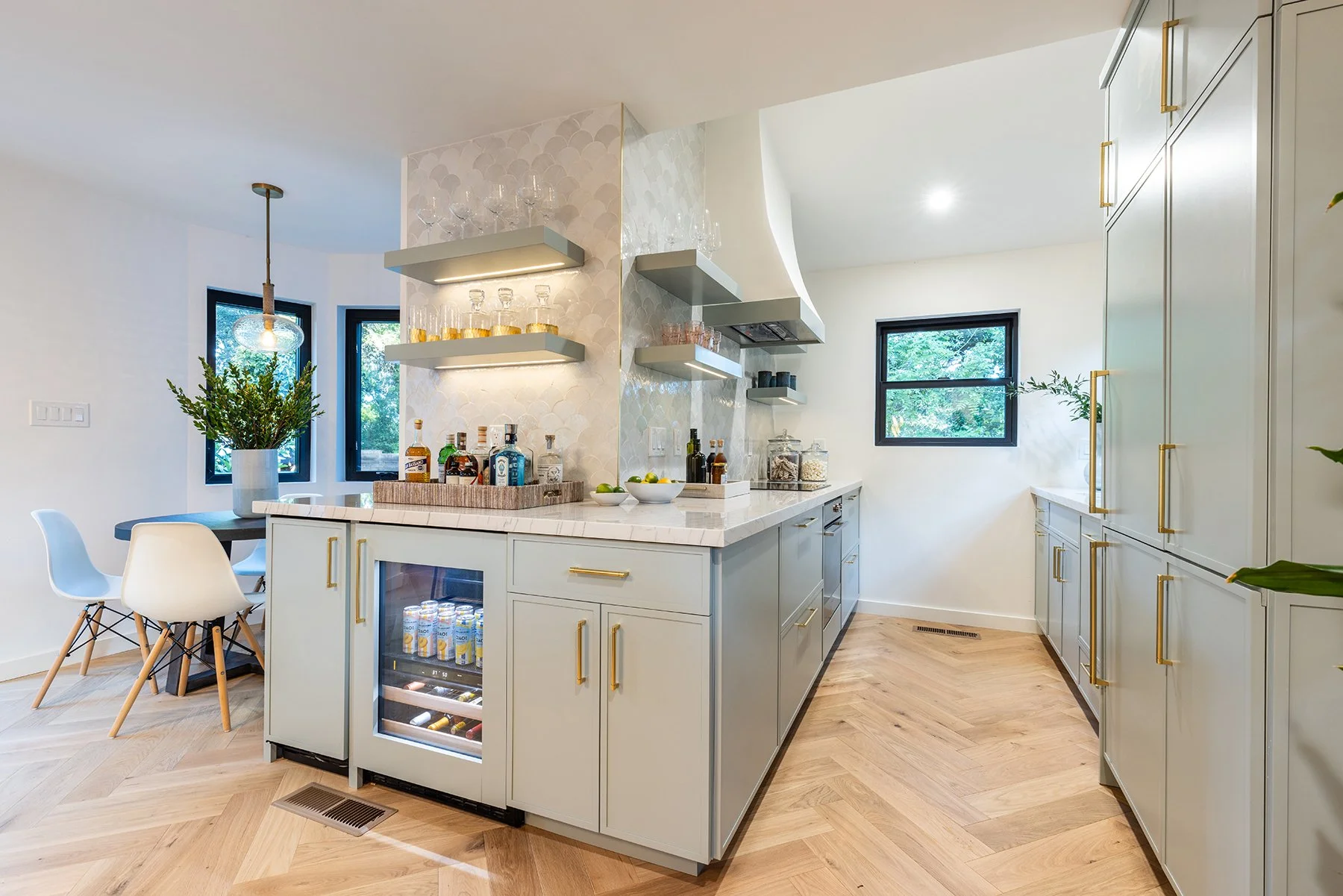 Modern kitchen with green cabinetry, gold hardware, marble island with bar stools, open shelving with glassware, small refrigerator, and large windows with greenery outside.