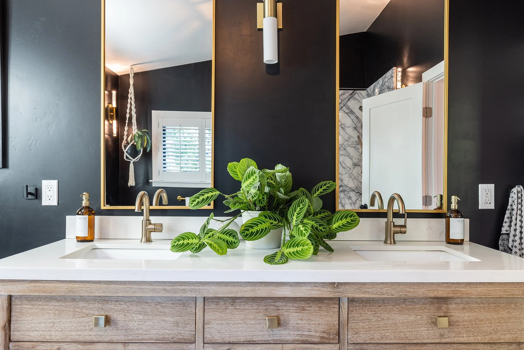 A modern bathroom vanity with a white countertop, two sinks, and brass fixtures. There are two amber soap dispensers, a potted green plant in the center, and two large mirrors with gold frames. The mirror on the left shows a window with white shutter