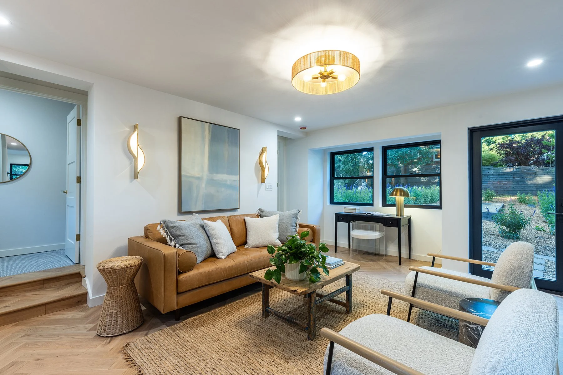 Living room with white walls, wooden flooring, tan leather sofa with white and gray pillows, a rustic wooden coffee table, a plant, two white armchairs with wooden armrests, a dark desk with a brass lamp, large windows with dark frames, and a door le