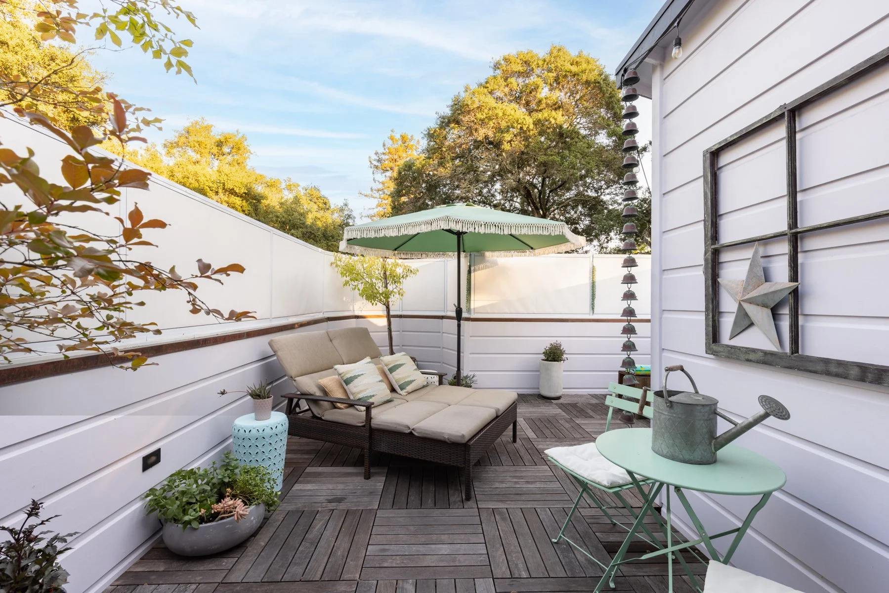 This is an outdoor patio with seating, a green patio umbrella, potted plants, and decorative items including a star on the wall, surrounded by white fencing and trees in the background.