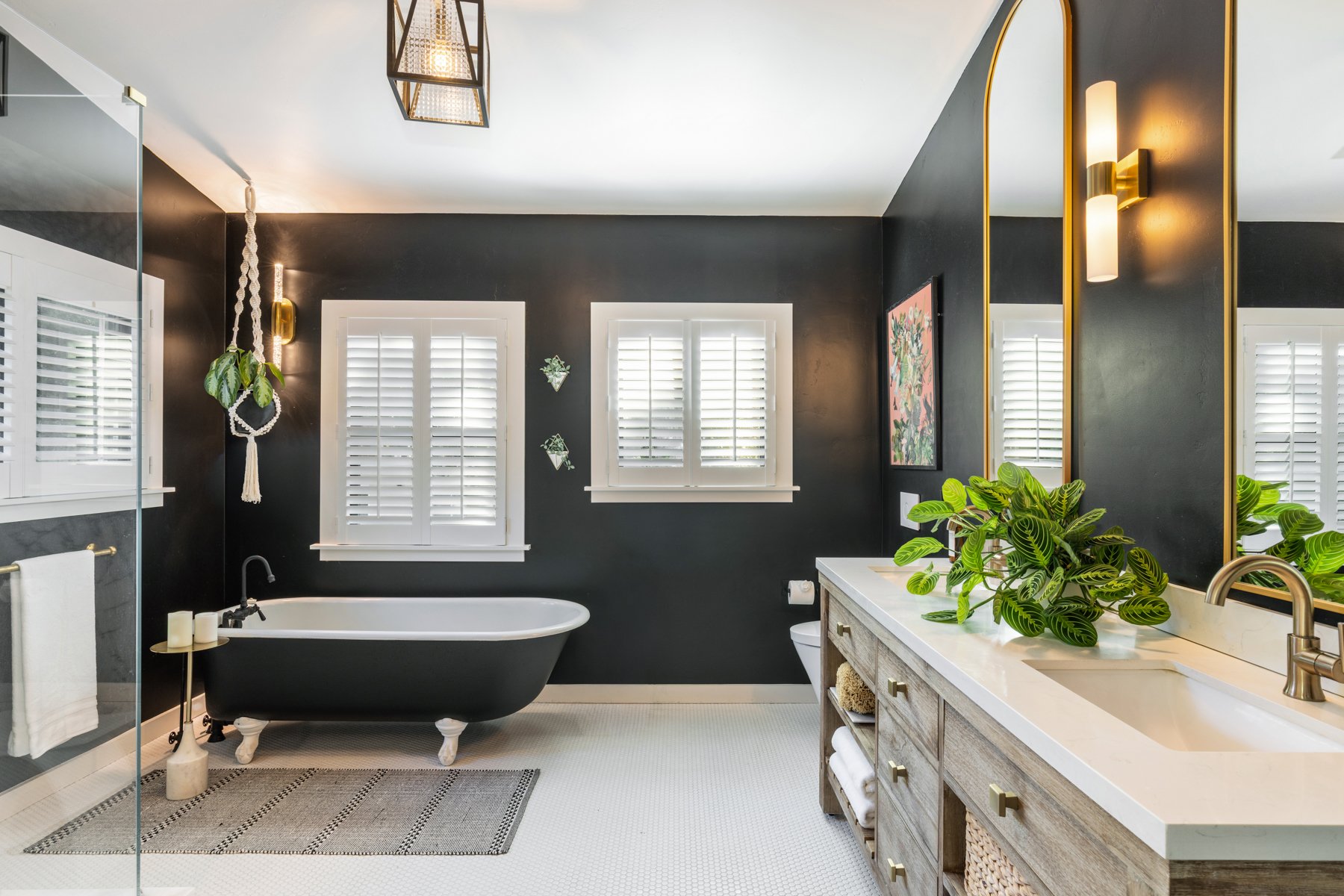 Modern bathroom with black walls, white shiplap window shutters, a black clawfoot bathtub with candles, a light grey vanity with a large mirror, green plants, and gold accents.