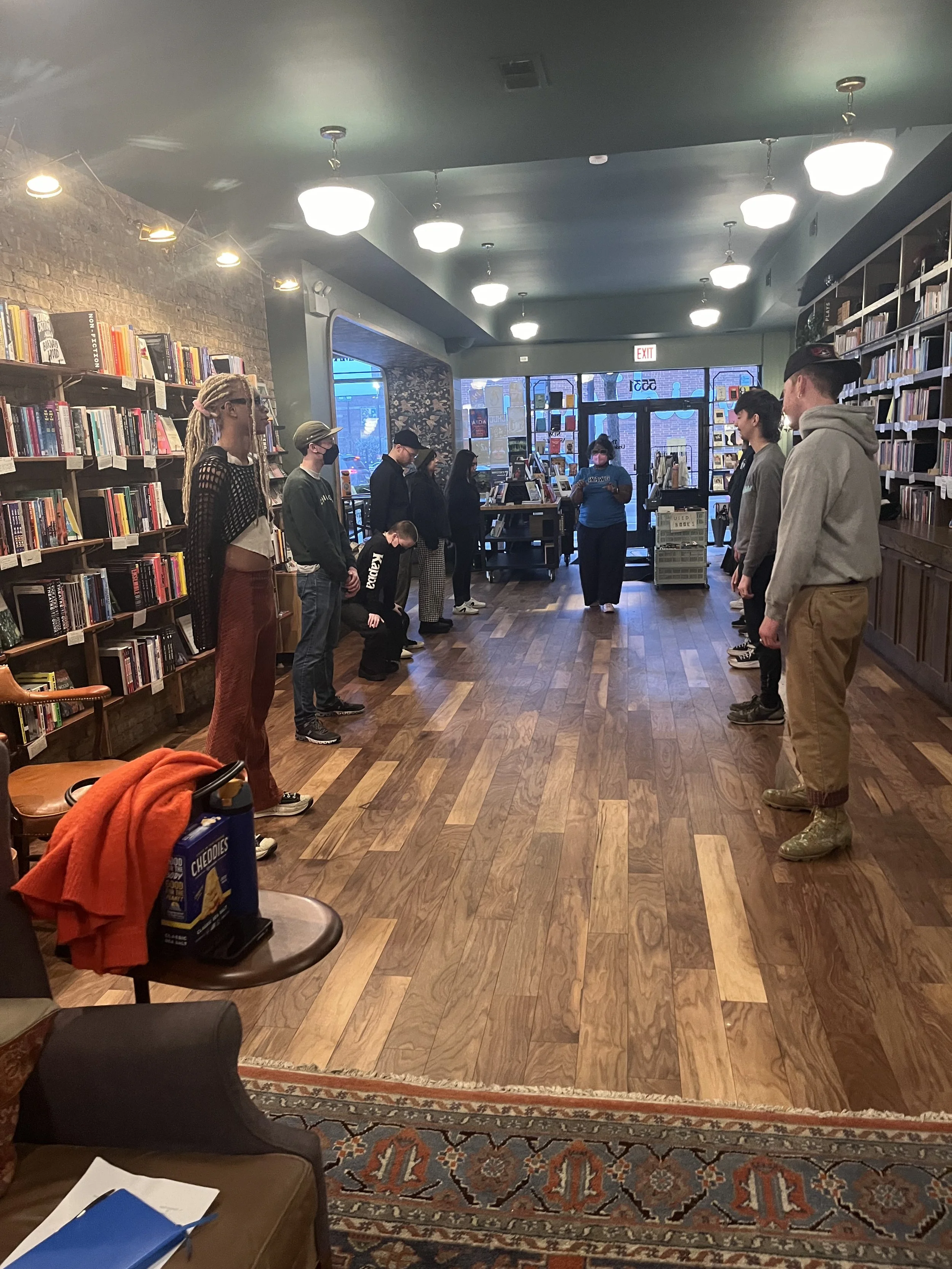 A group of people standing in a circle inside a bookstore or library, with bookshelves lining the walls. Some people are wearing masks.