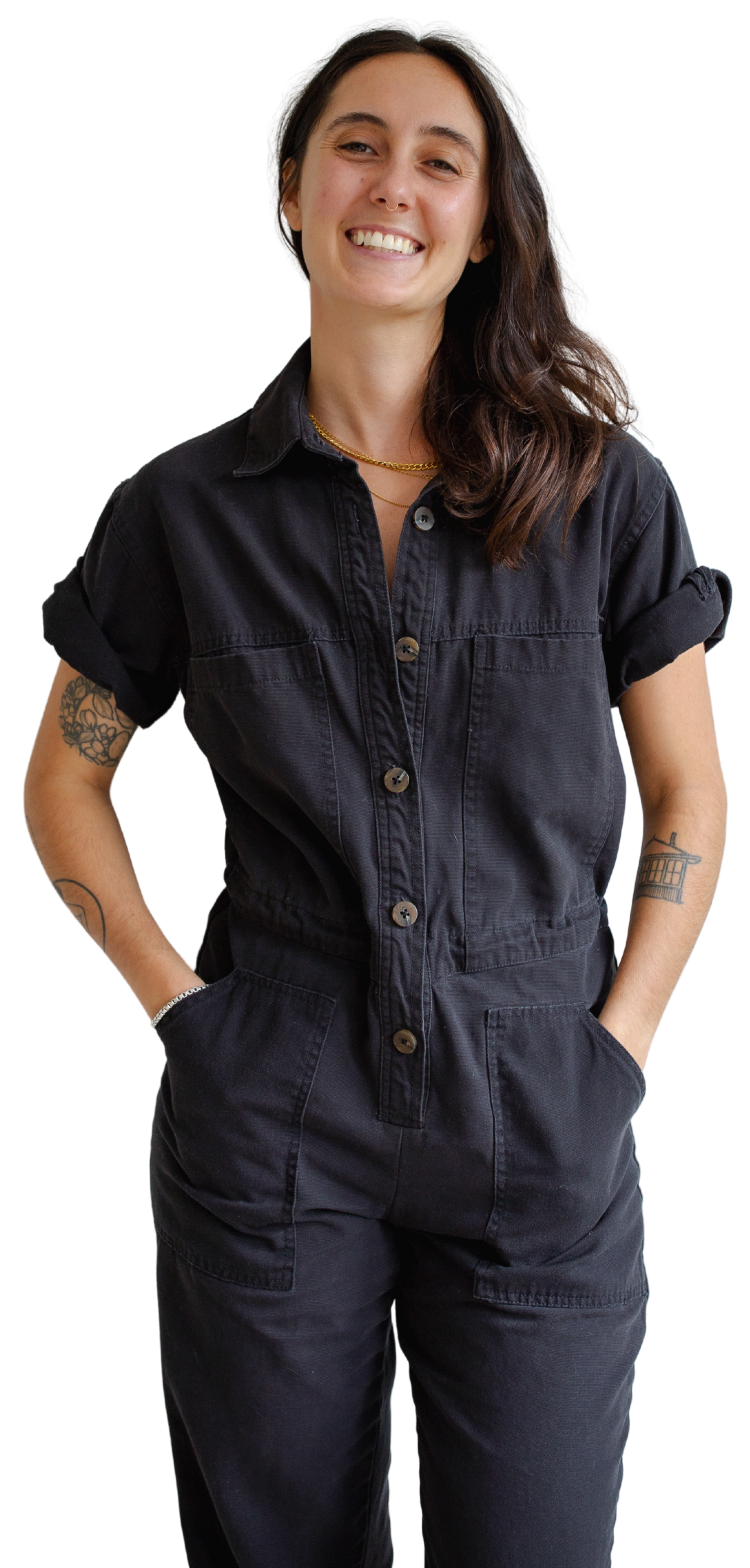 Kate stands with their hands in their pockets, wearing a black denim jumpsuit with rolled-up sleeves, smiling.